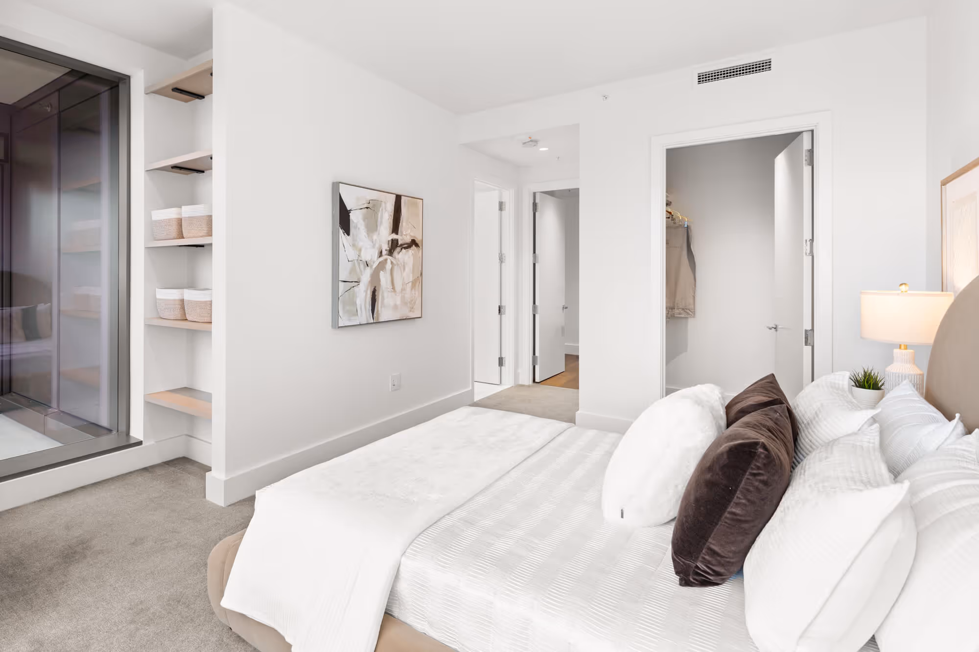 Modern bedroom  at The Ritz-Carlton Residences, Portland with a neatly made bed featuring white and dark brown pillows, a bedside lamp on a nightstand, an abstract painting on a white wall, and an open closet door.