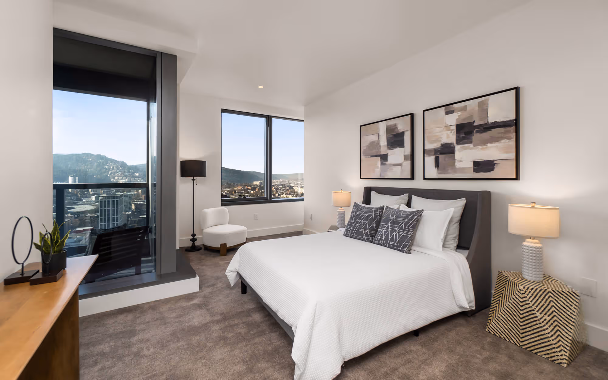 Modern bedroom at The Ritz-Carlton Residences, Portland with white bed linens, abstract art above the bed, a white chair by the window, and Portland skyline view through large windows.