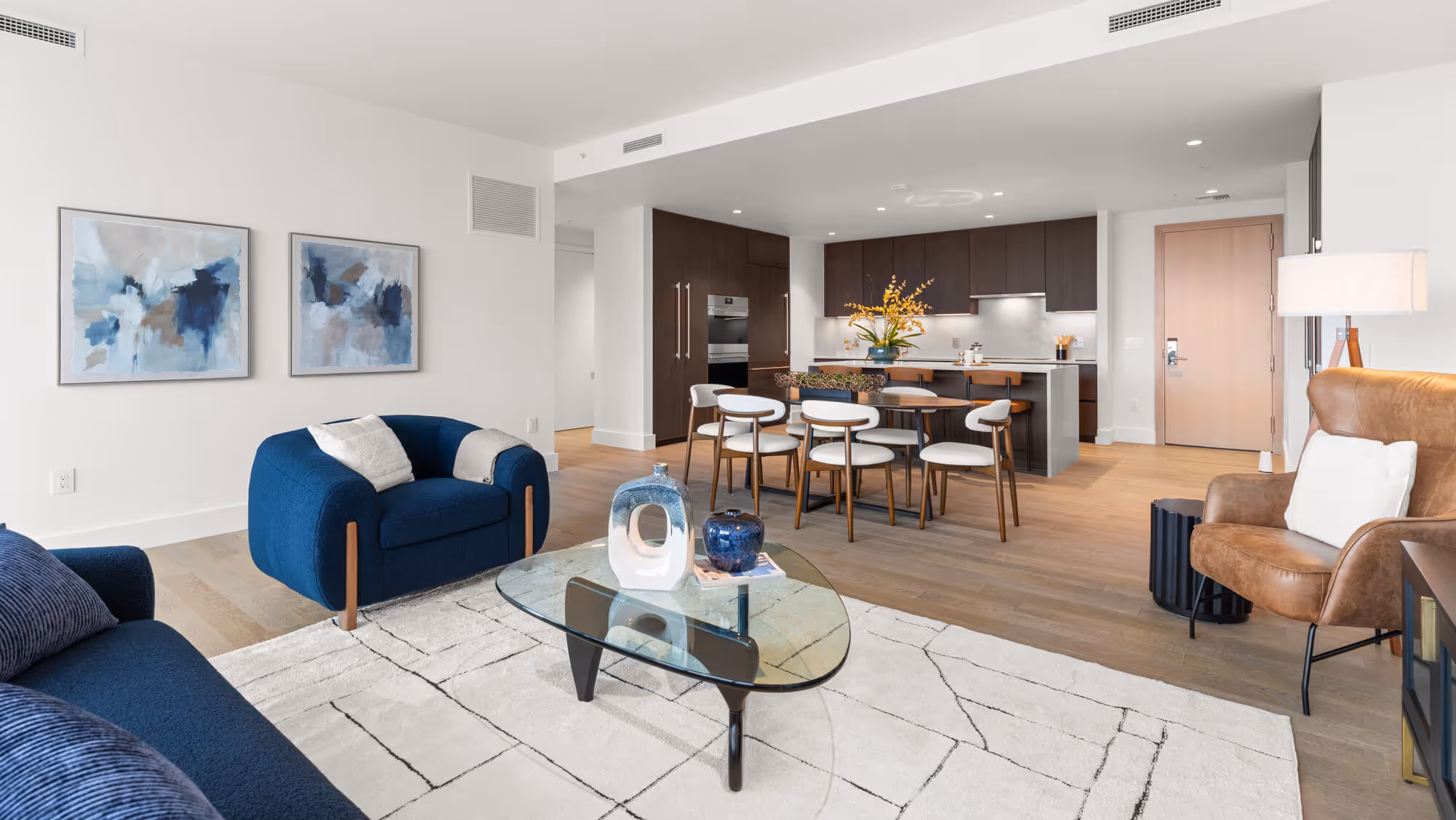 Modern living room at The Ritz-Carlton Residences, Portland with blue armchairs and sofa, glass coffee table on white patterned rug, and dining area with wooden chairs and dark kitchen cabinets in the background.