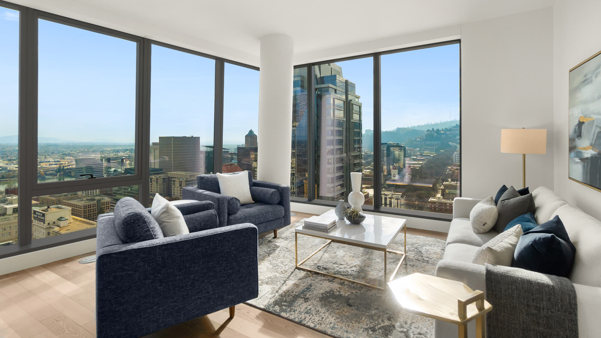 Modern living room at The Ritz-Carlton Residences, Portland with large floor-to-ceiling windows showing the Portland skyline, featuring blue armchairs, a white couch with assorted pillows, a coffee table, and a standing lamp.
