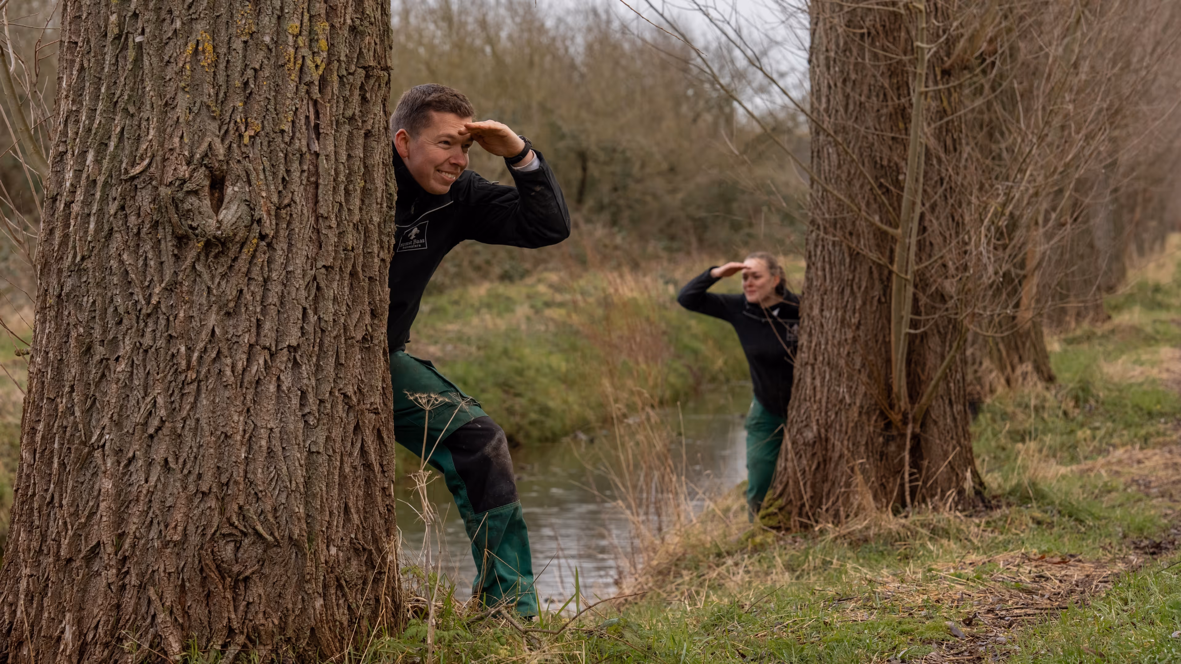 Two people in work clothes playfully peeking from behind trees near a narrow stream in a wooded area.