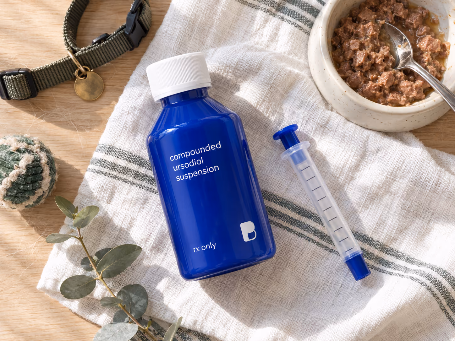 Flatlay of compounded ursodiol suspension with an oral syringe and a bowl of pet food, prepared by Bayview Pharmacy