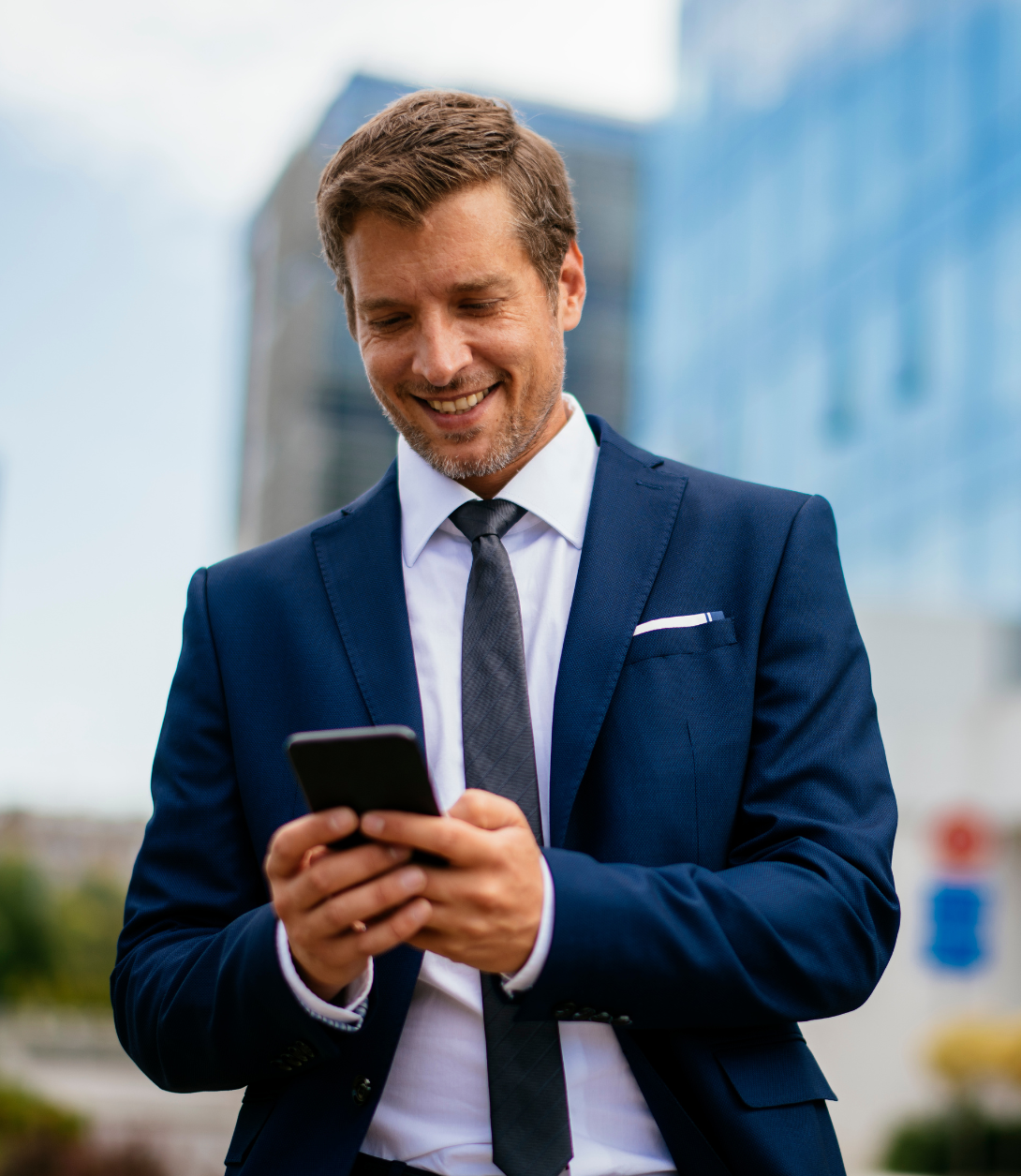 Geschäftsmann im blauen Anzug, der lächelnd auf sein Smartphone schaut, mit modernen Gebäuden im Hintergrund.
