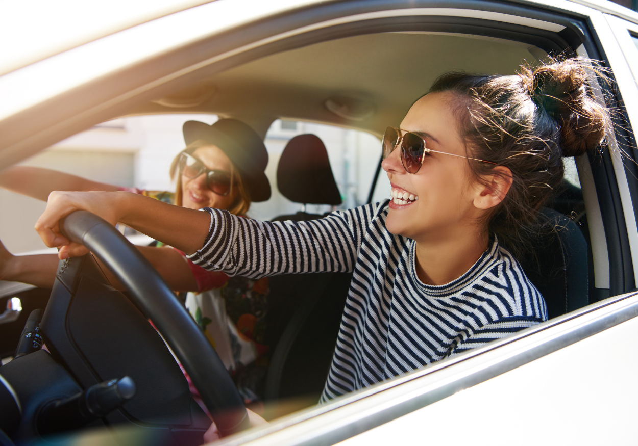 Zwei lächelnde Frauen mit Sonnenbrillen sitzen in einem Auto, die Fahrerin hält das Lenkrad, während die Beifahrerin einen Hut trägt.