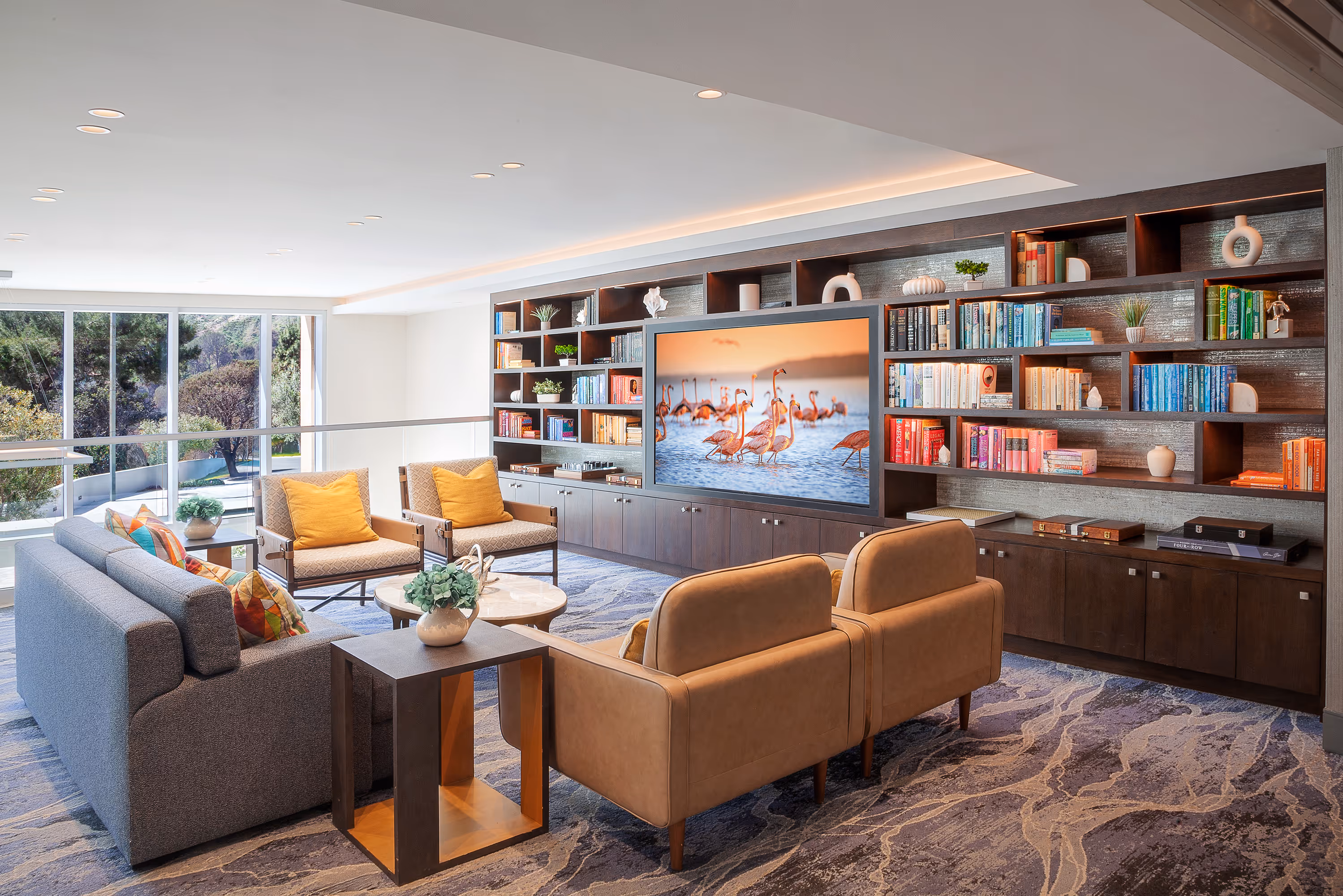 Modern living room with a large built-in bookshelf filled with colorful books, a flat-screen TV displaying flamingos, and various seating including a gray sofa, two tan armchairs, and two patterned chairs with yellow pillows.