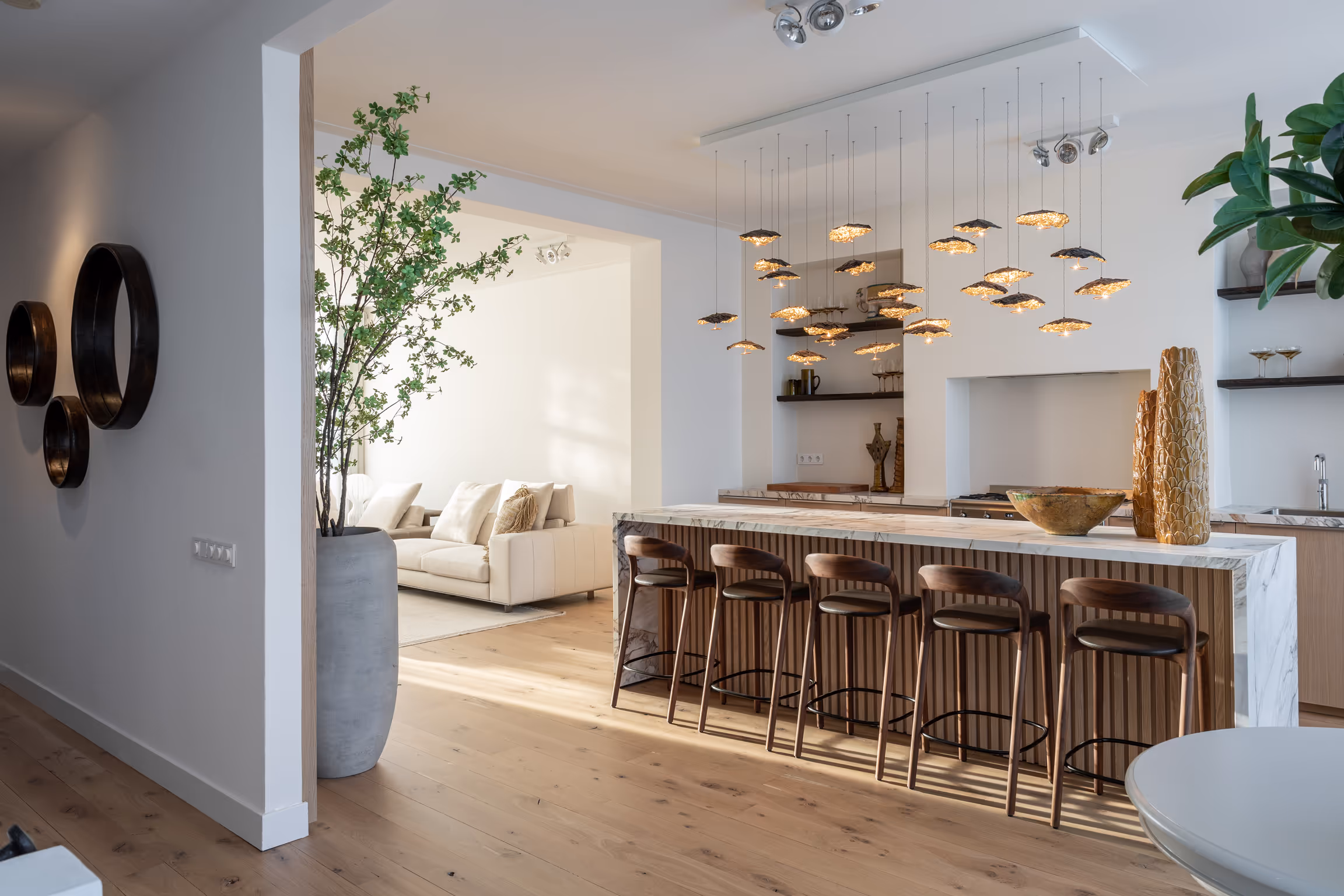 Moderne open keuken met marmeren kookeiland, vijf houten barkrukken, hangende pendellampen en aangrenzende woonkamer met witte bank.