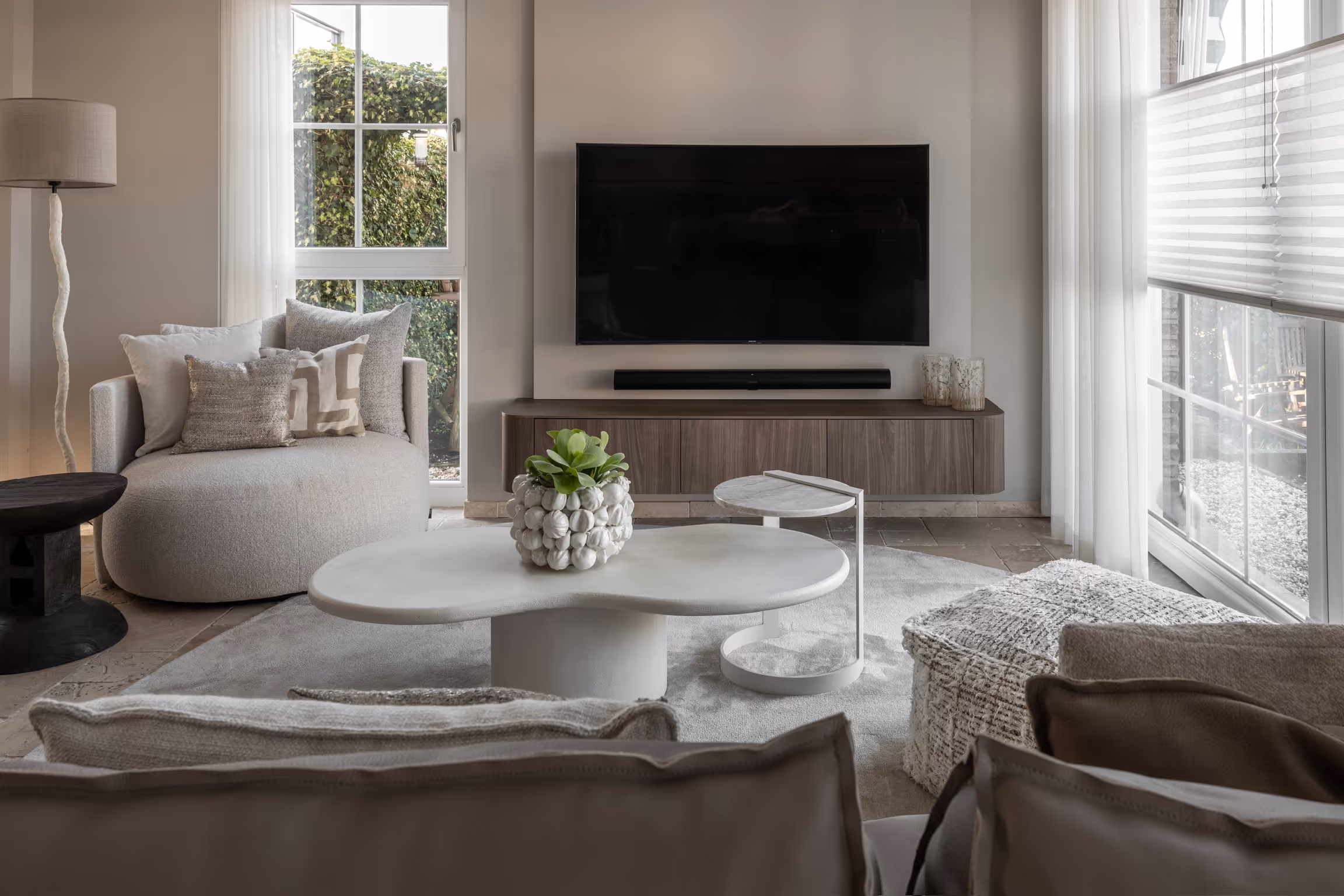 Moderne woonkamer met beige fauteuil, witte salontafel met een plant, aan de muur bevestigde tv en grote ramen met witte gordijnen.