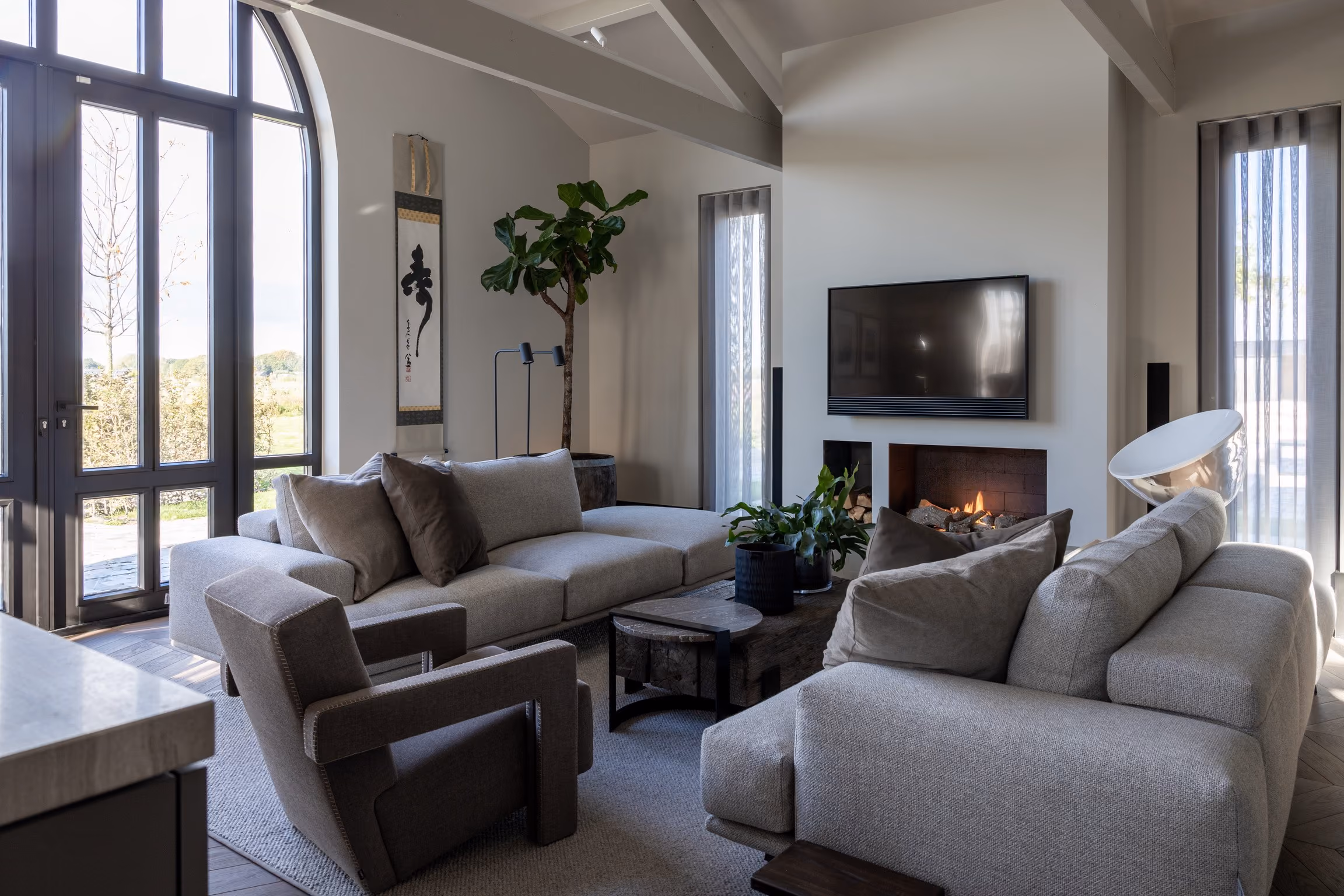 Modern living room with grey sofas, armchair, wooden coffee table, indoor plants, wall-mounted TV, and a lit fireplace.