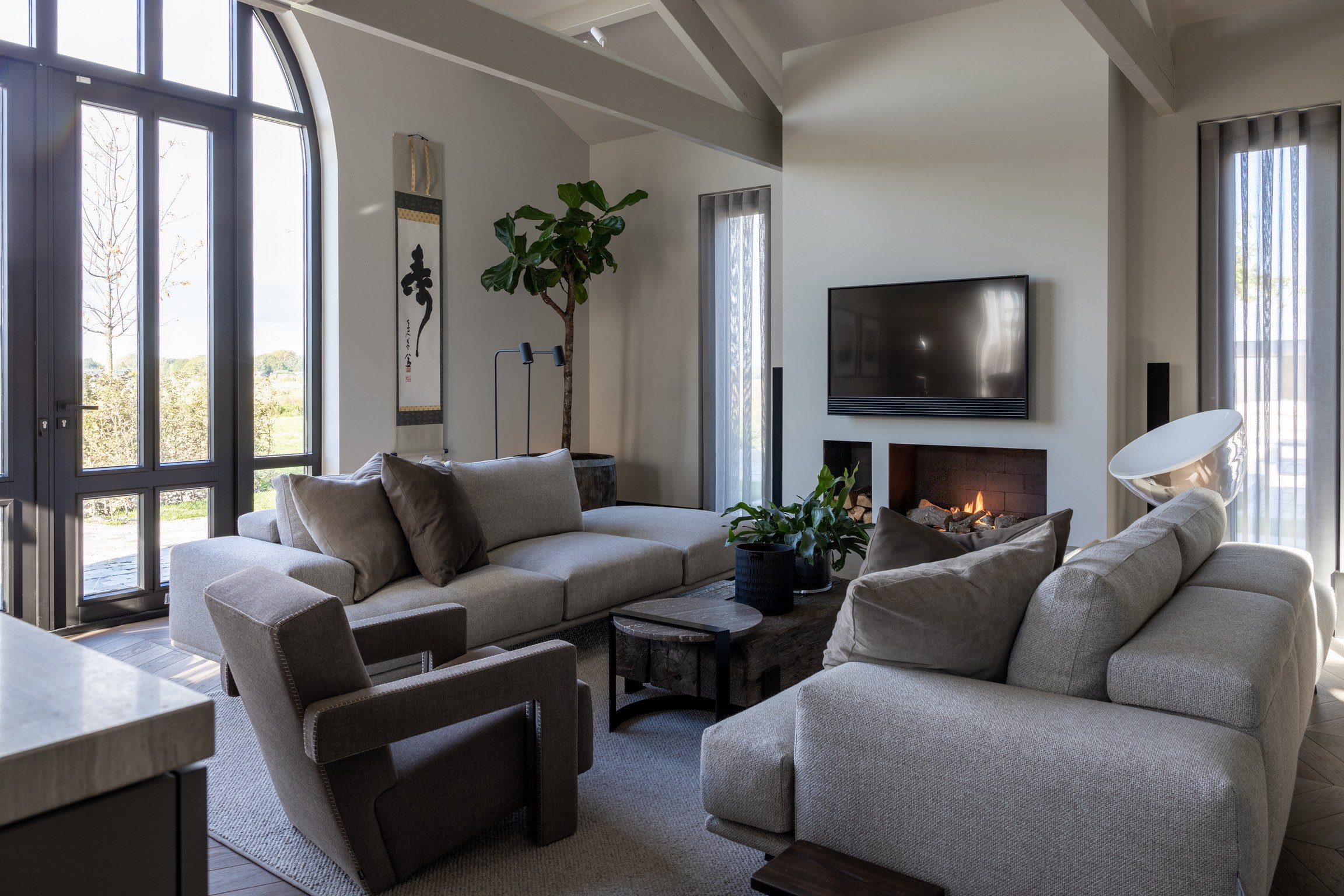 Moderne woonkamer met grijze banken, fauteuil, houten salontafel, kamerplanten, aan de muur bevestigde tv en een brandende open haard.