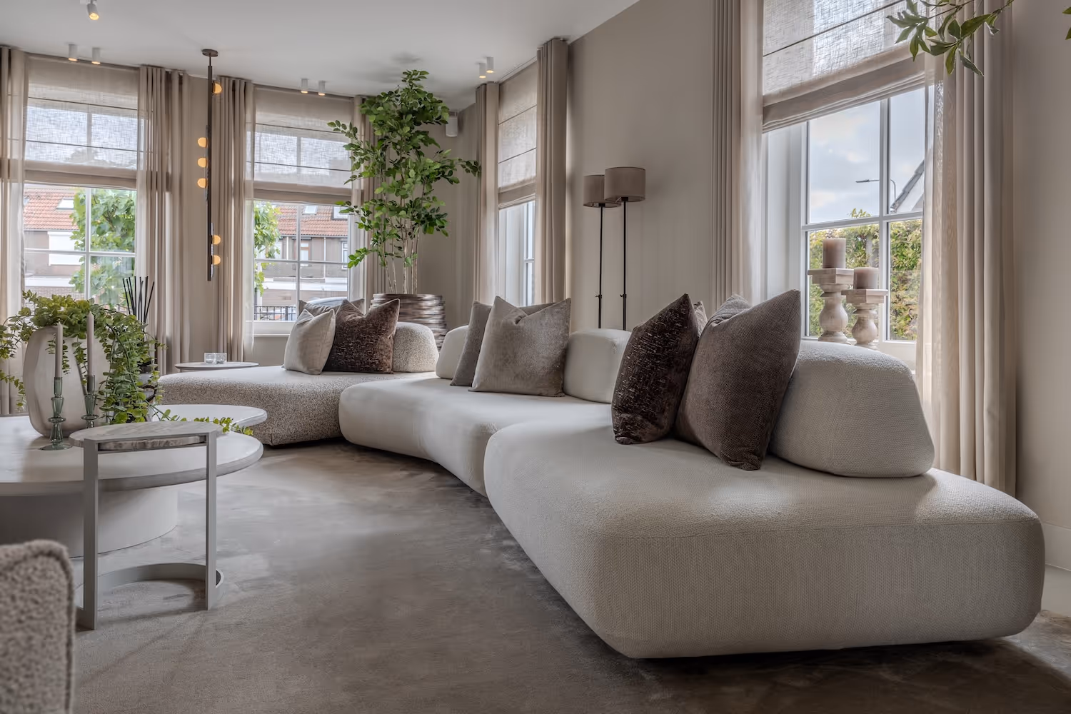 Moderne woonkamer met grote, gebogen witte bank versierd met grijze en bruine kussens, grote ramen met beige gordijnen en kamerplanten.