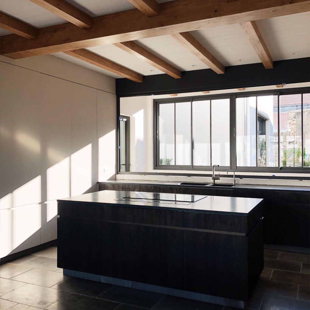 Modern kitchen with dark island countertop, stovetop, large window, and exposed wooden ceiling beams.