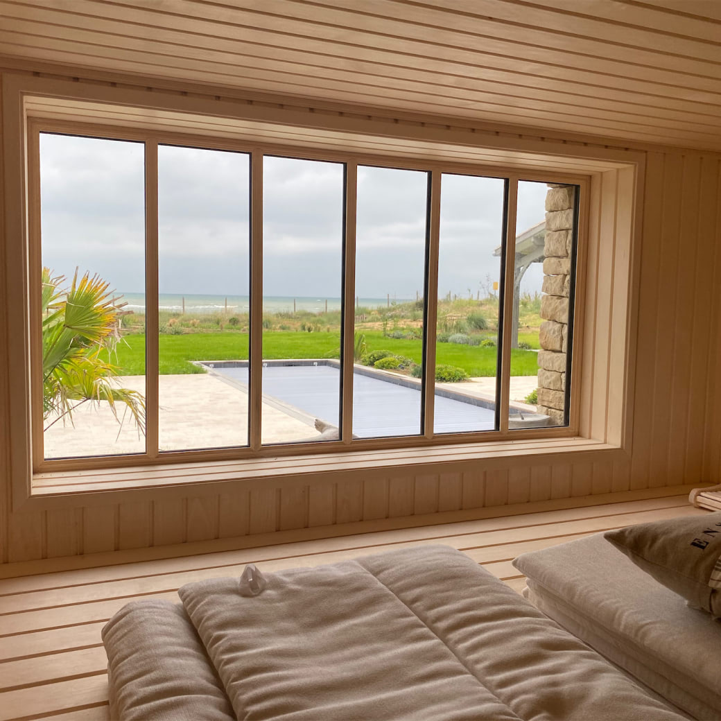 View through large wooden-framed window of a garden with a covered pool, green lawn, and an ocean horizon under a cloudy sky.