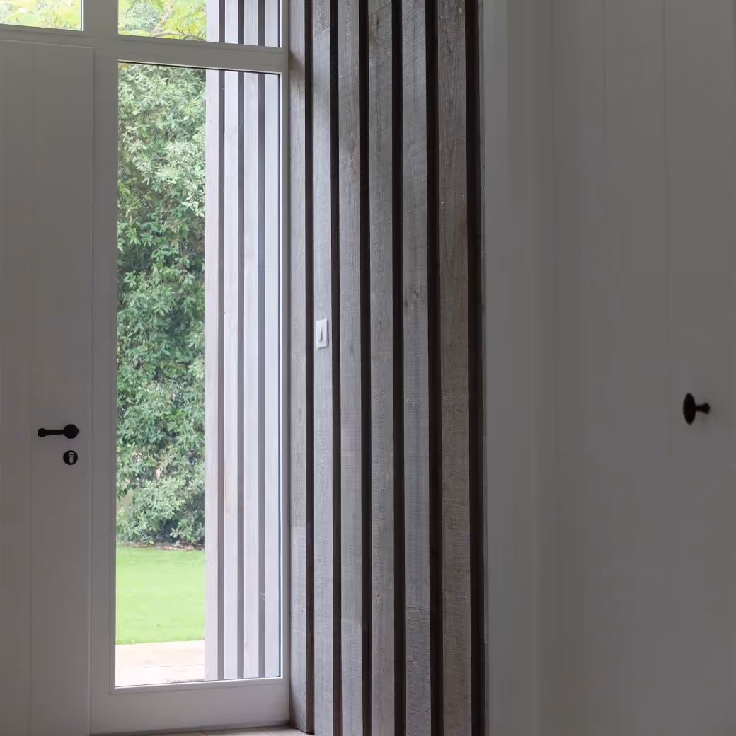Interior view of a modern doorway with a glass panel showing green foliage and grass outside, flanked by vertical wooden slats and white walls with black door handles.