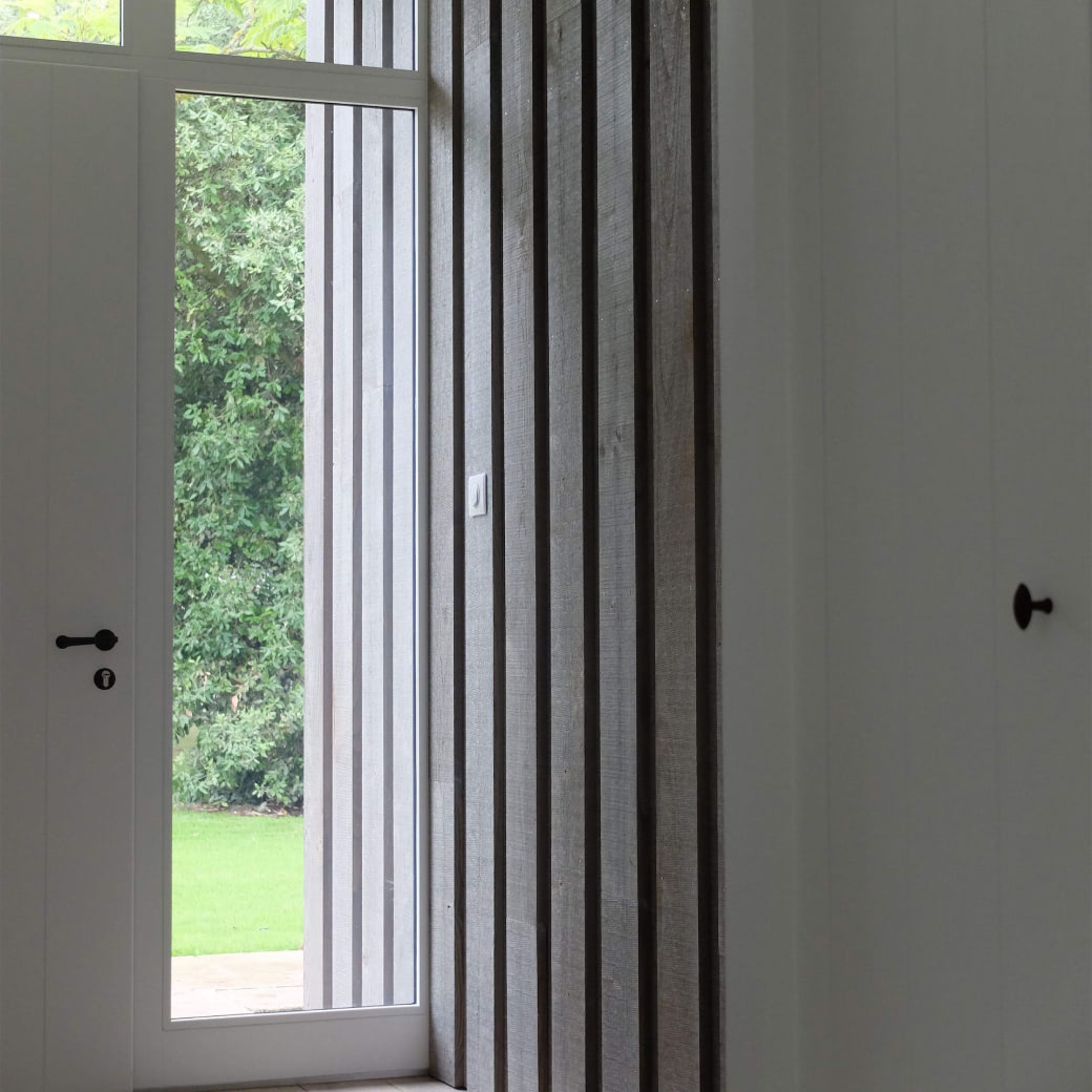 Interior view of a modern doorway with a glass panel showing green foliage and grass outside, flanked by vertical wooden slats and white walls with black door handles.