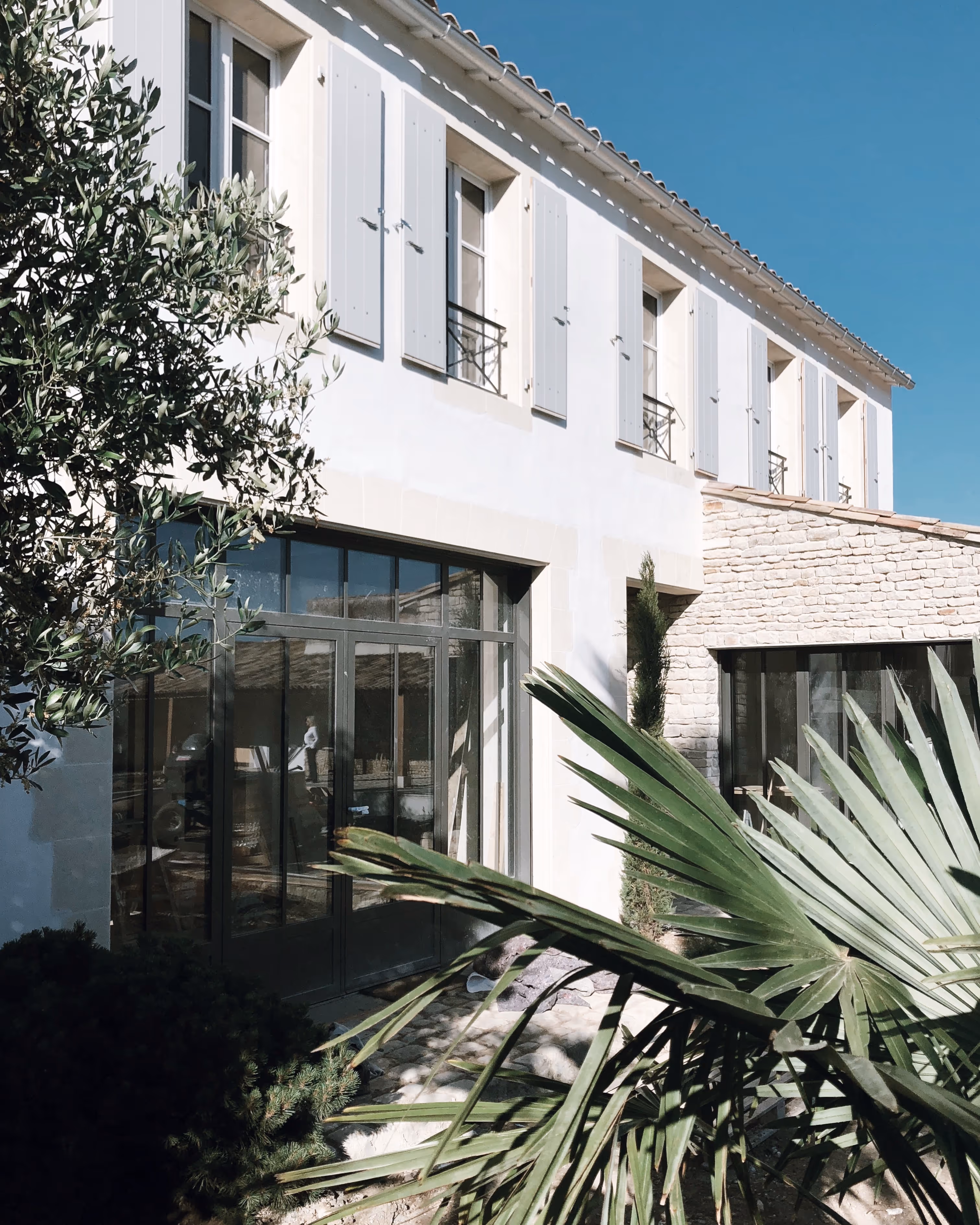 Modern two-story house with white shutters, large glass doors, stone wall section, and greenery in the foreground.