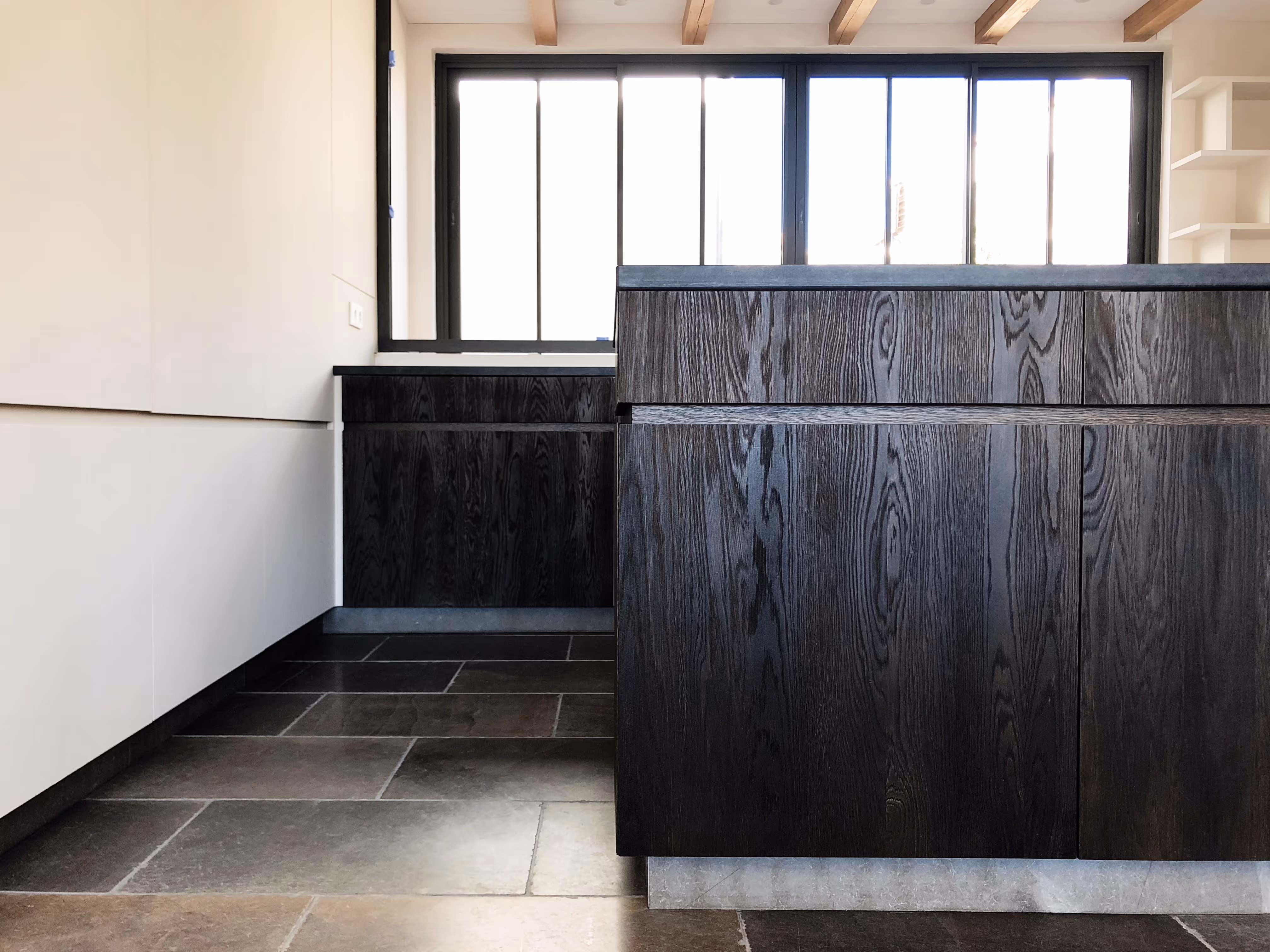 Modern kitchen interior with dark wood cabinets, white walls, large window, and stone tile floor.