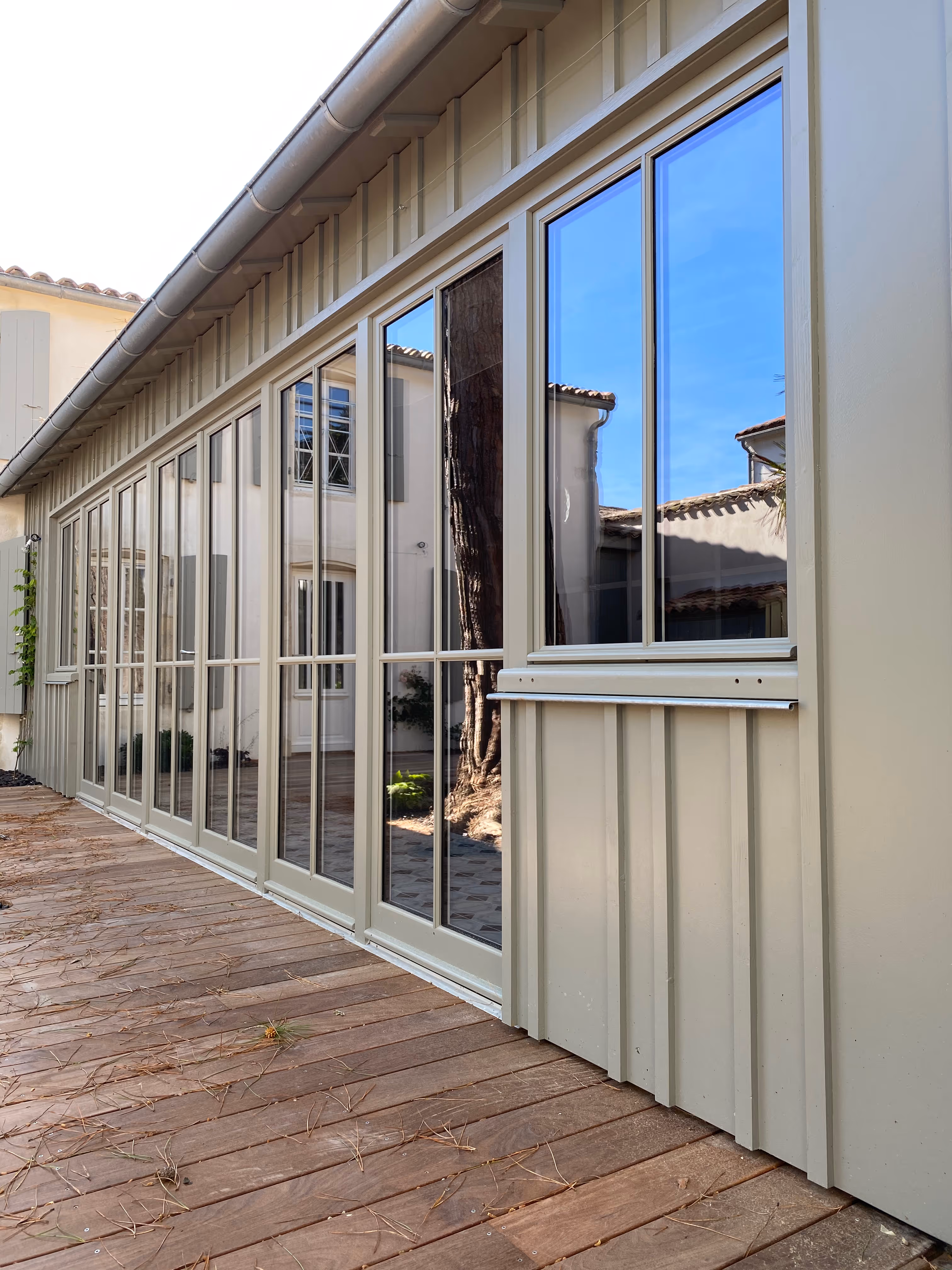 View of a beige house exterior with large glass windows reflecting blue sky and surrounding buildings, next to a wooden deck scattered with pine needles.
