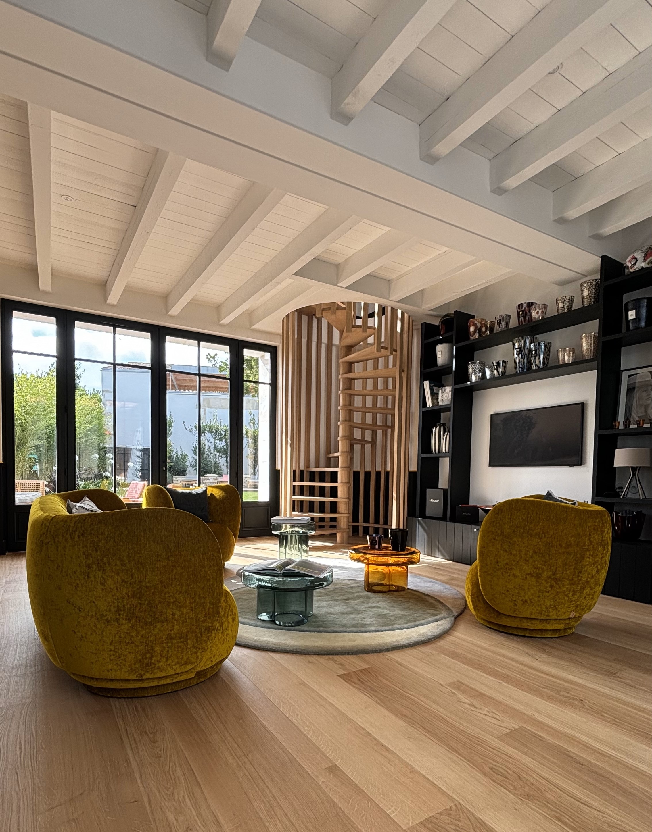 Modern living room with three mustard yellow velvet chairs around a round rug and glass coffee tables, featuring a wooden spiral staircase and large windows.