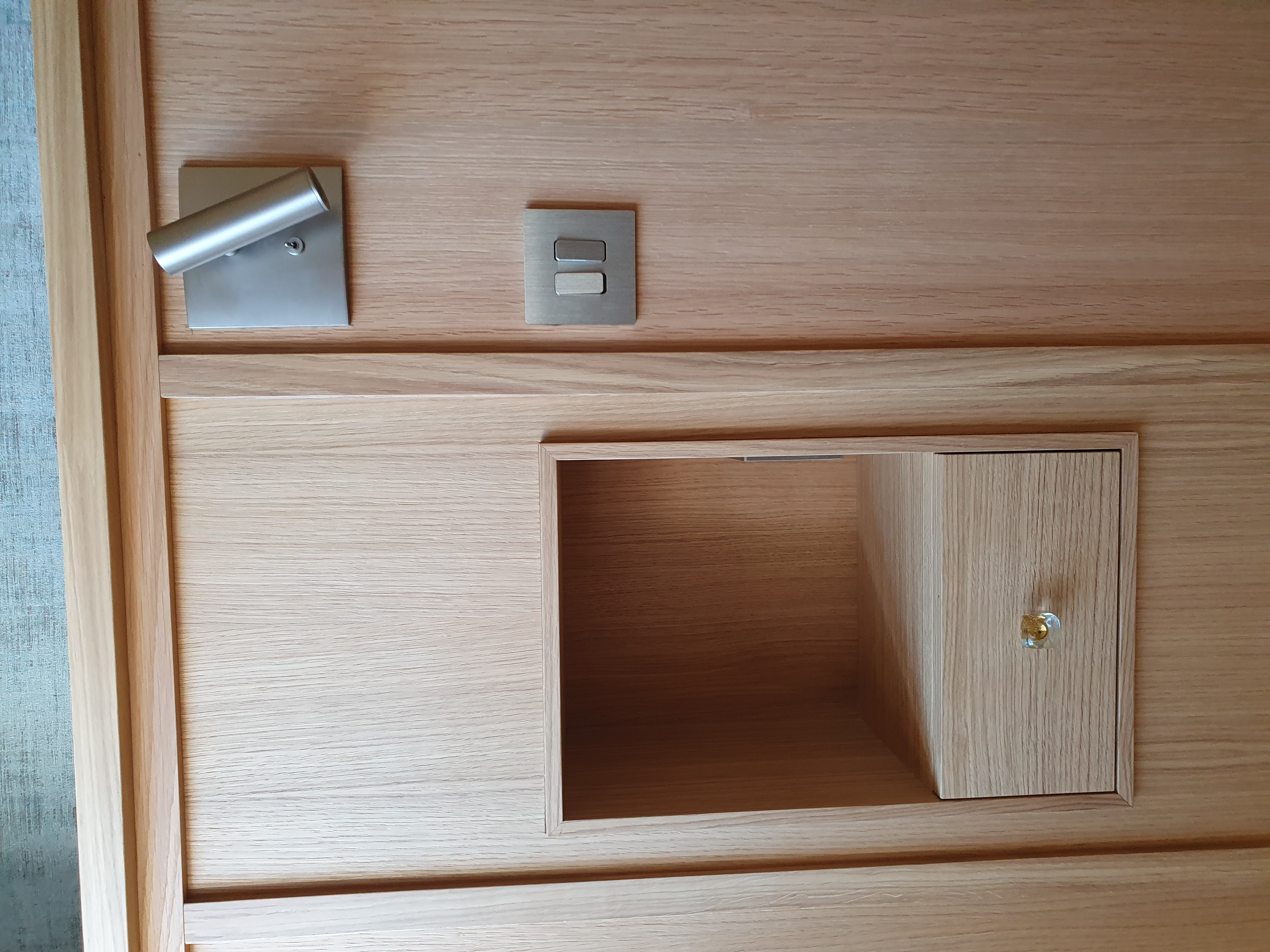 Wooden panel with a small open shelf including a drawer with a glass knob, a metal spotlight above, and a double light switch.