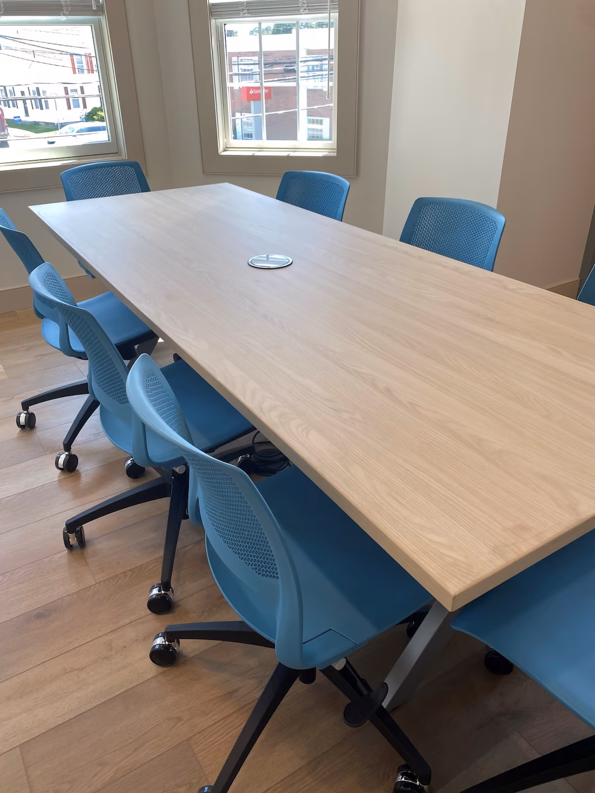 Modern conference room with light wood table and ergonomic blue chairs on hardwood floor, featuring large windows and natural lighting