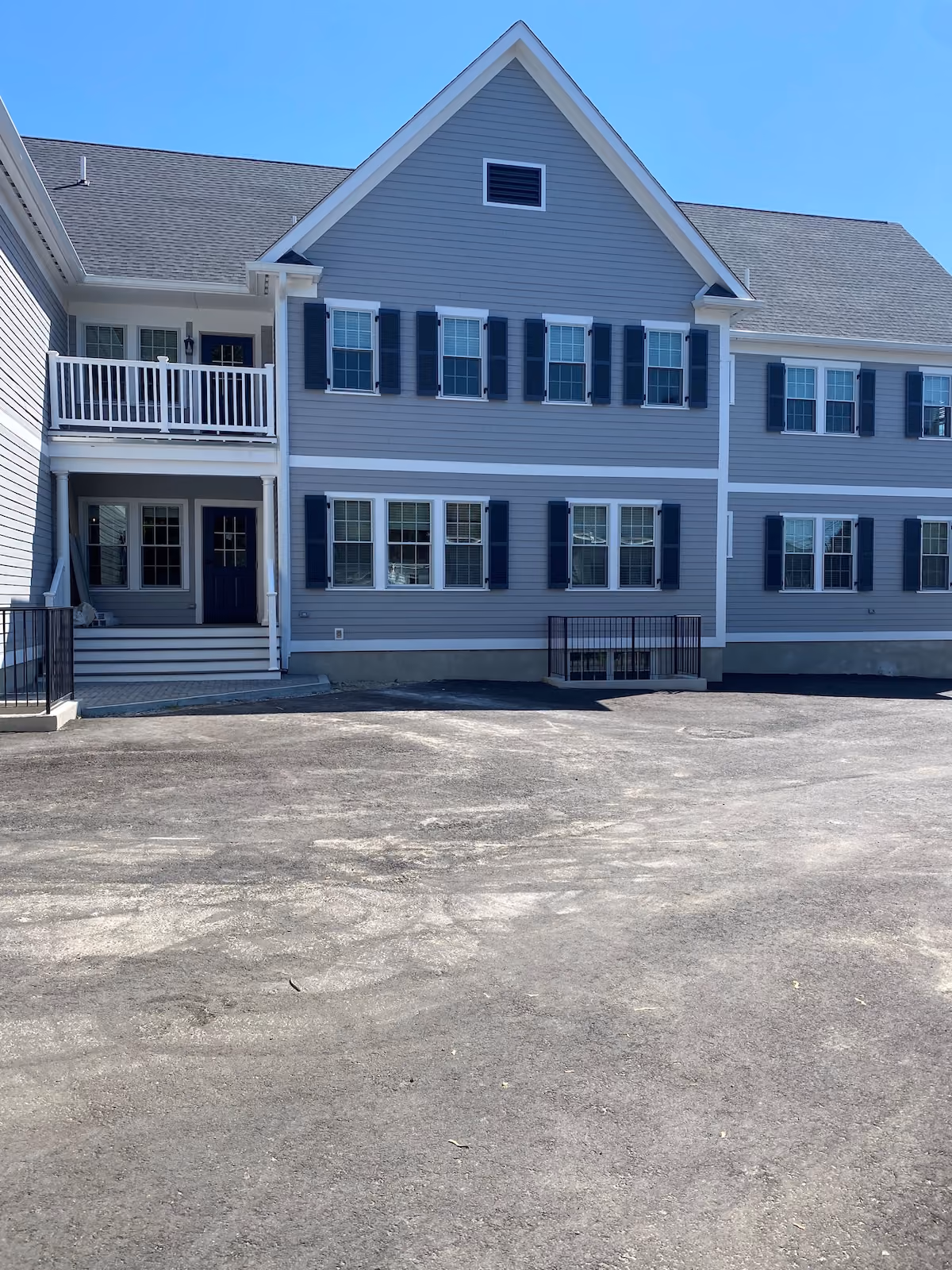Two-story gray medical or office building with navy shutters and large parking area under clear blue sky