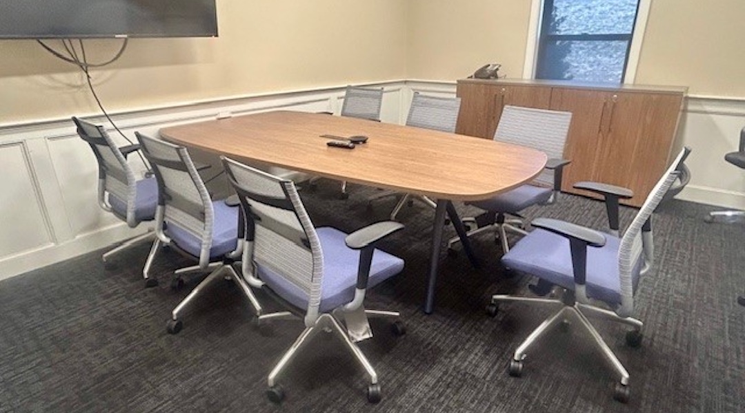Rectangular wood-grain conference table with eight ergonomic rolling chairs in a fully equipped meeting room with mounted monitor