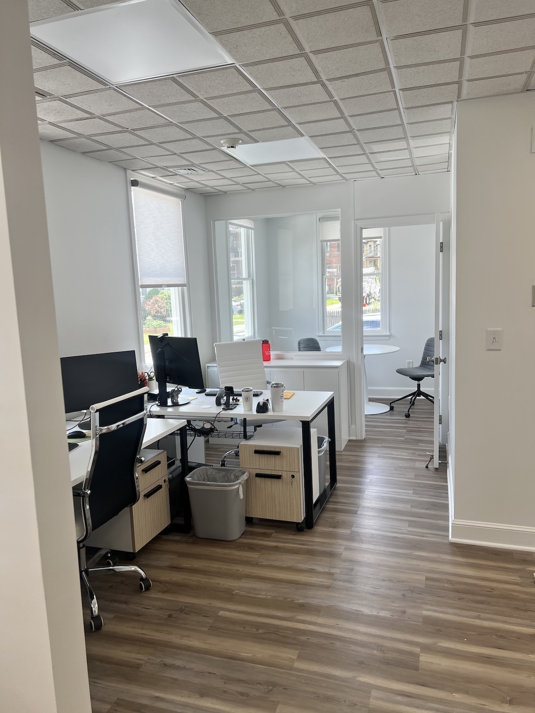 Bright real estate corporate office interior featuring open-plan workstations with white desks, ergonomic black and white office chairs, dual monitor setups, and wood-look vinyl plank flooring under a grid ceiling.