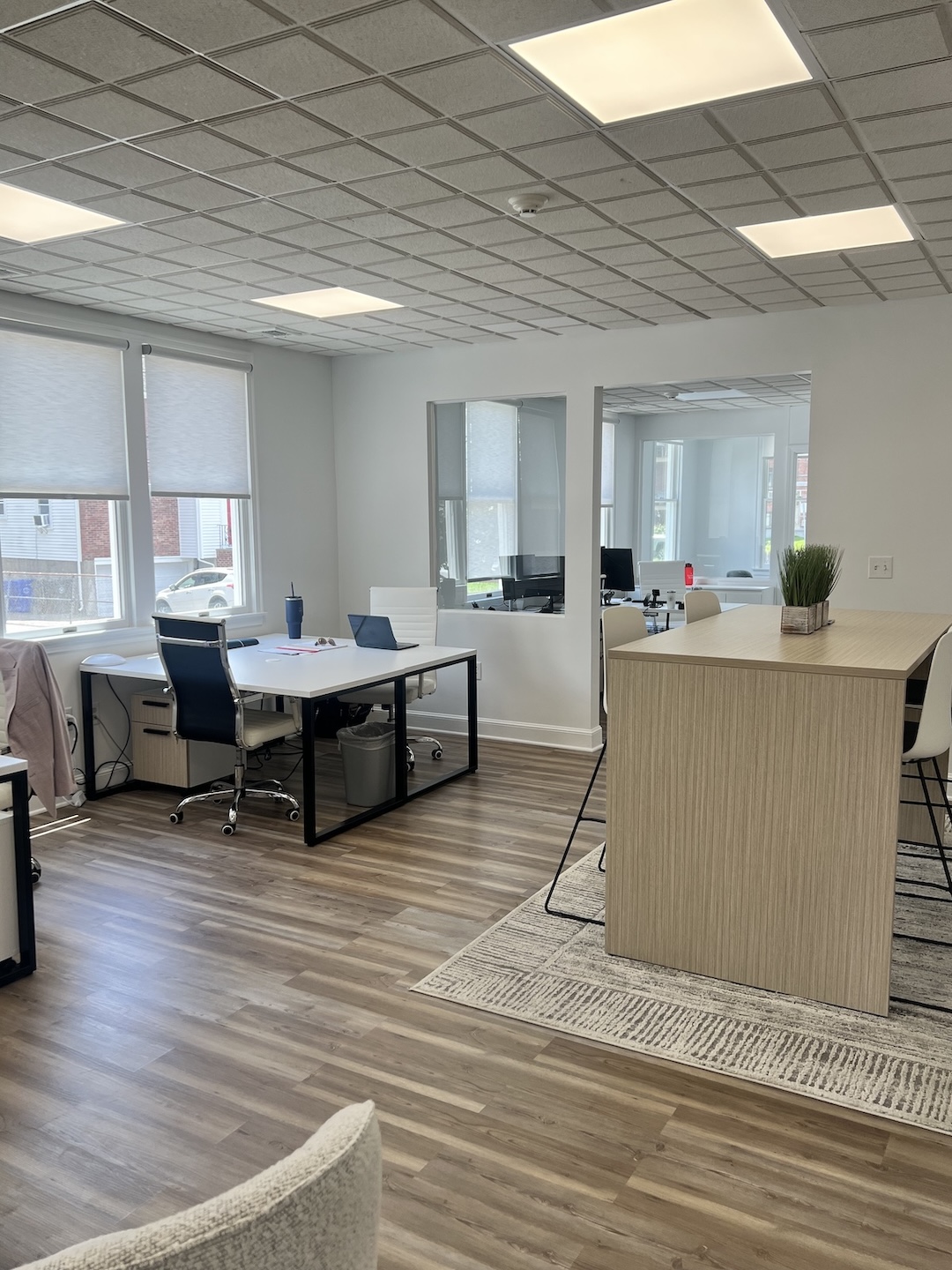 Modern office interior with dual desk workstations, white ergonomic chairs, and a light wood standing-height collaboration table placed over a geometric area rug next to large windows.