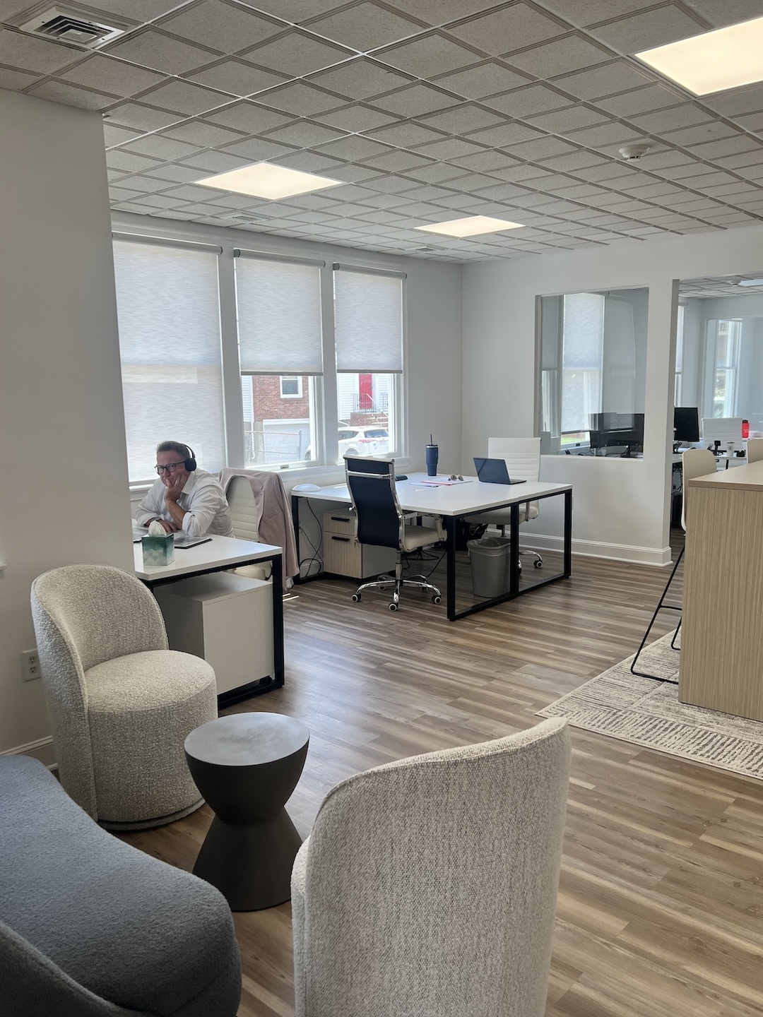 Open office workspace view displaying desk pods with a professional working at a laptop, a high-top collaborative table, and a cozy client waiting area with textured grey accent chairs.