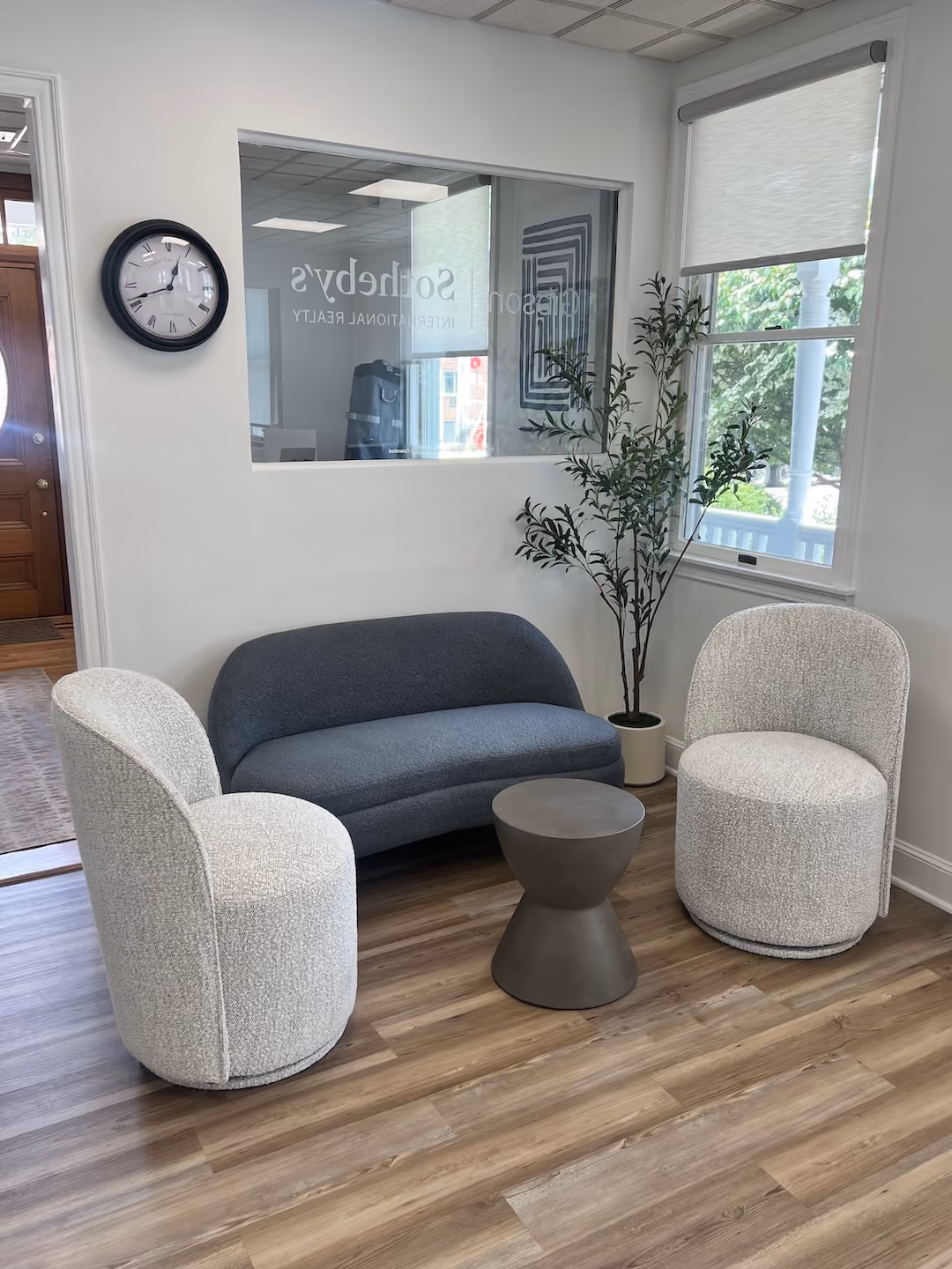 Gibson Sotheby's International Realty office lobby waiting area featuring a dark grey boucle sofa, two cream textured accent chairs, a concrete barrel side table, and a large wall clock.