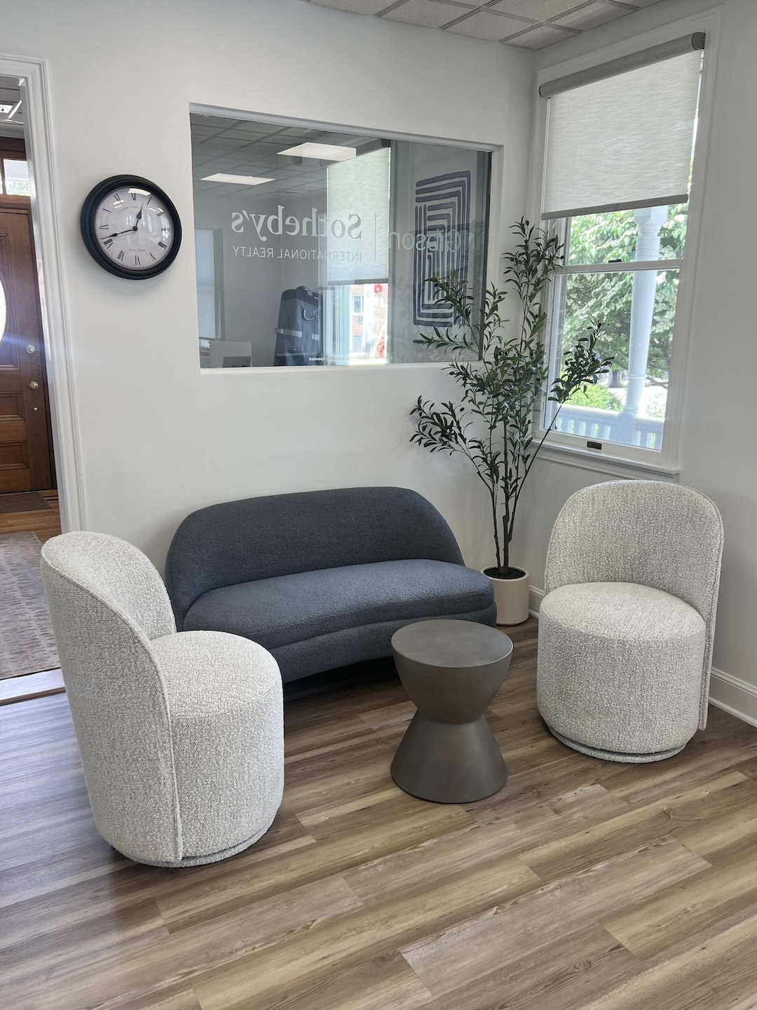 Gibson Sotheby's International Realty office lobby waiting area featuring a dark grey boucle sofa, two cream textured accent chairs, a concrete barrel side table, and a large wall clock.