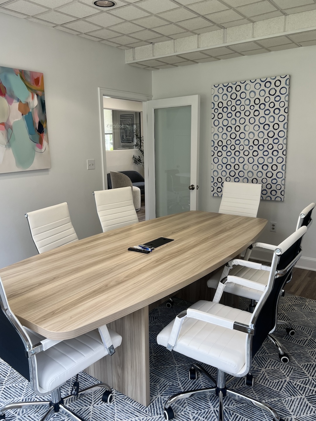 Professional conference room featuring a light oak wood boardroom table, modern white leather ribbed chairs, abstract wall art, and a blue geometric patterned rug.