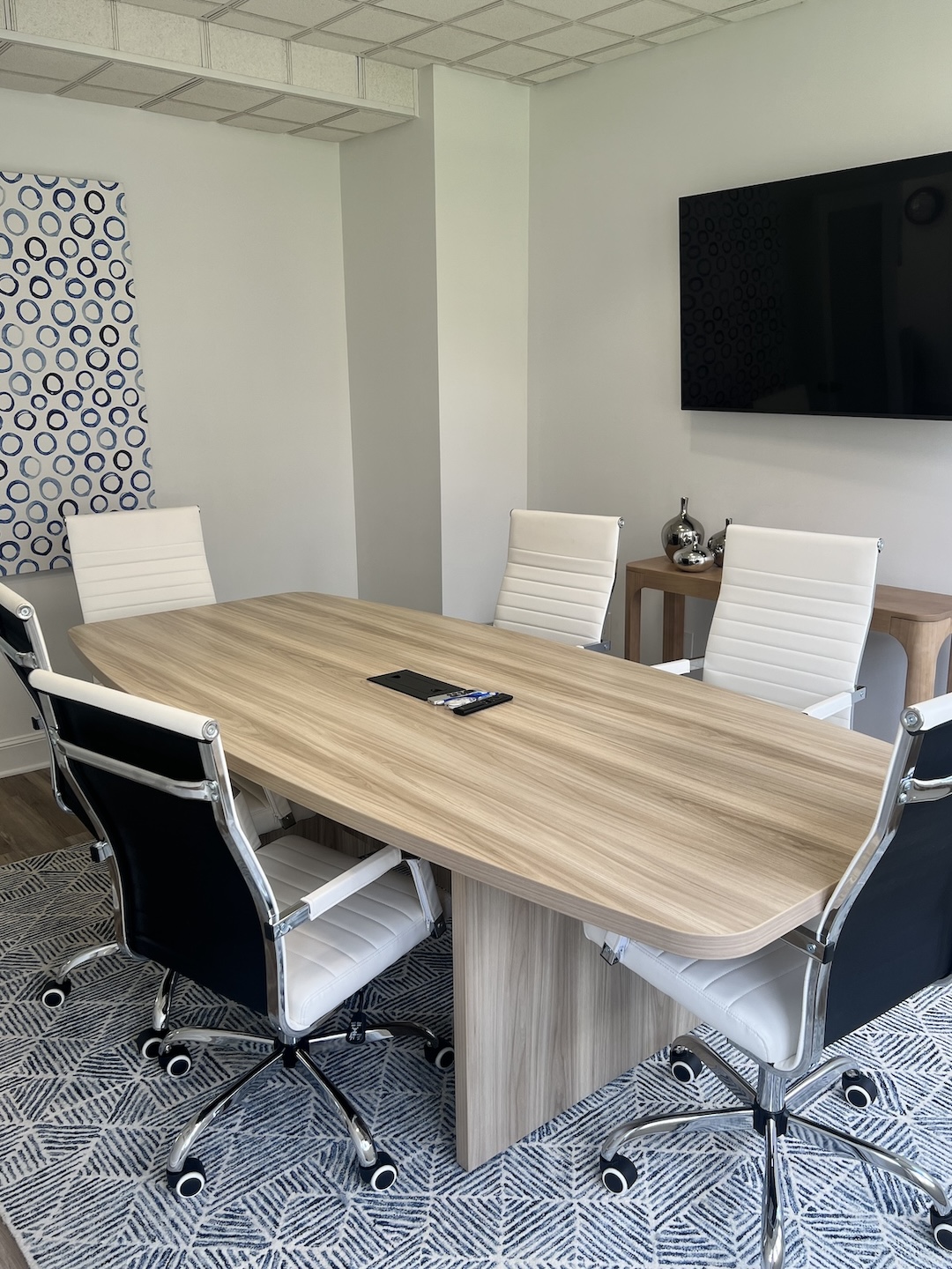 Luxury boardroom interior displaying a light wood conference table surrounded by white ribbed executive chairs, a wall-mounted flat-screen TV, and a wood console table with decorative silver vases.