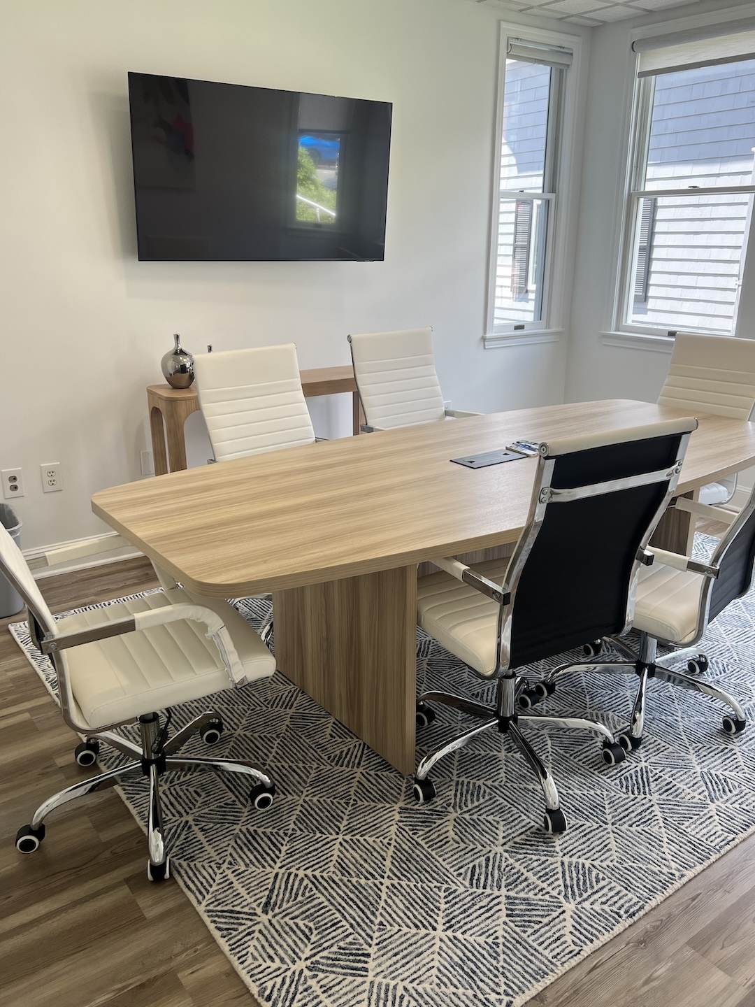Bright office conference room setup showing a long wooden meeting table, white leather rolling chairs, a wall-mounted display monitor, and large windows bringing in natural light.