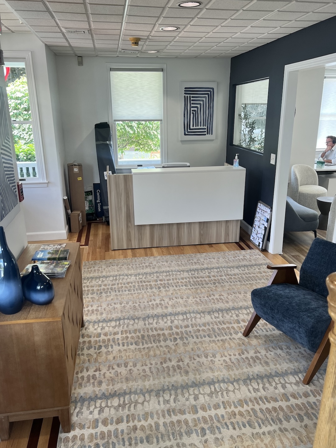 Real estate office reception lobby showcasing a modern white and wood reception desk, patterned area rug, blue mid-century accent chair, and a dark accent wall with a glass viewing window.