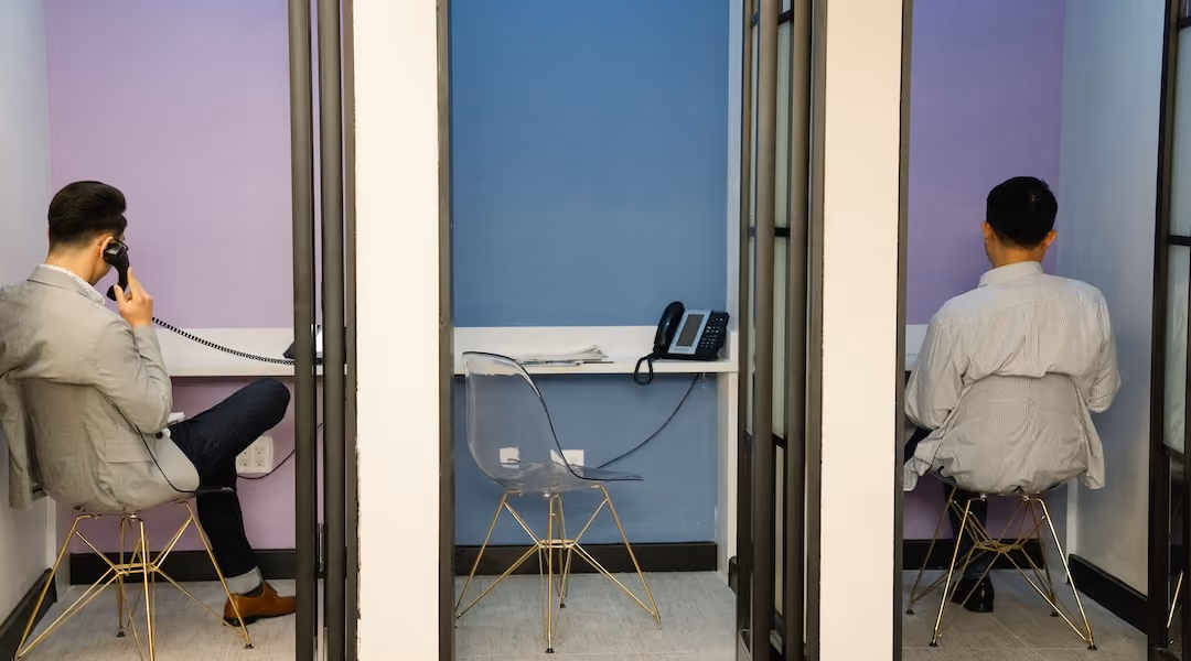 Two businessmen sit in contemporary phone booths using telephones in an office setting.