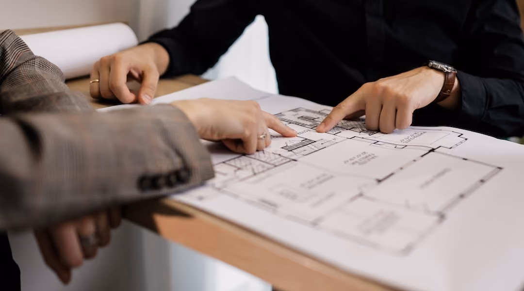 Two architects reviewing and pointing at a detailed floor plan on paper in an office setting.
