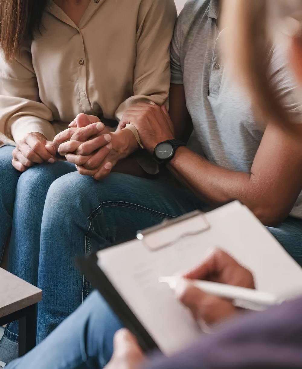 Man sitting on a sofa, appearing stressed, with hands clasped, in a counseling session.