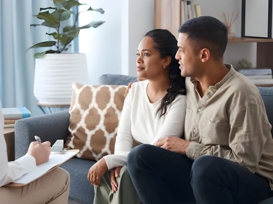 Man sitting on a sofa, appearing stressed, with hands clasped, in a counseling session.