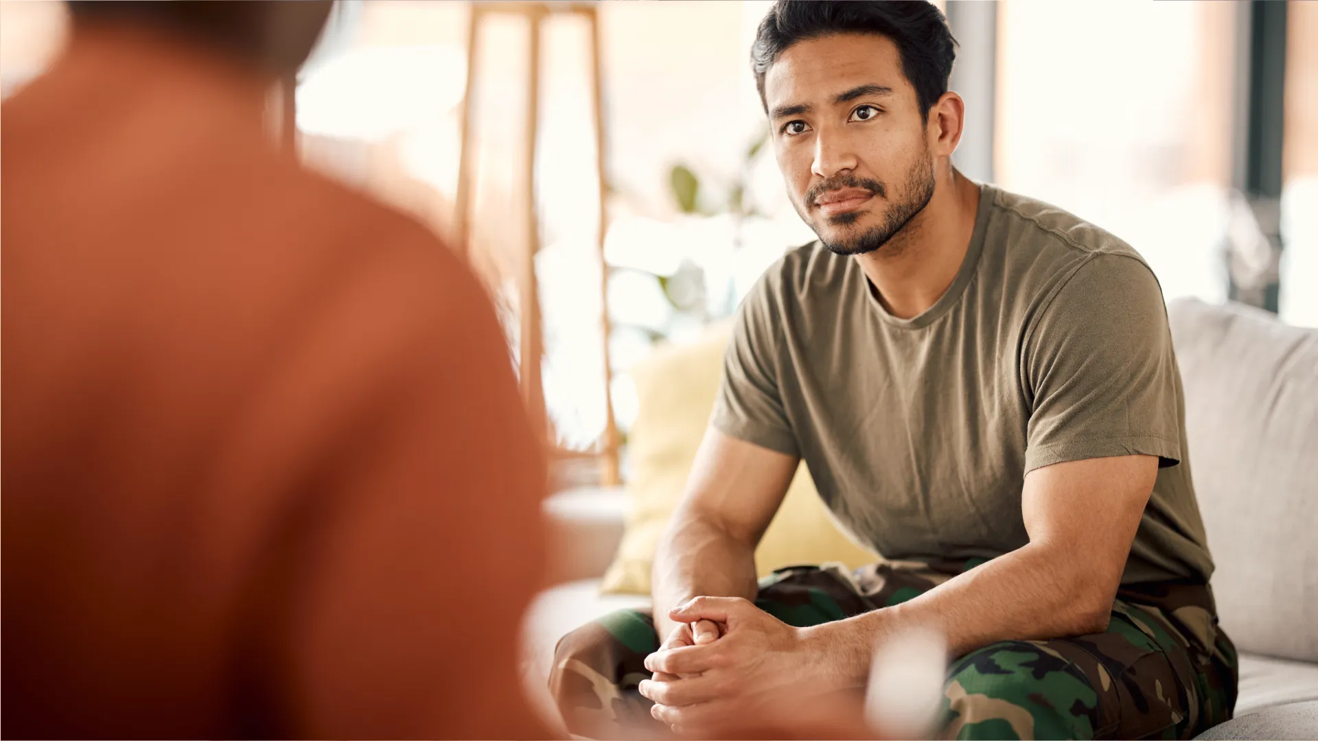 Man sitting on a sofa, appearing stressed, with hands clasped, in a counseling session.