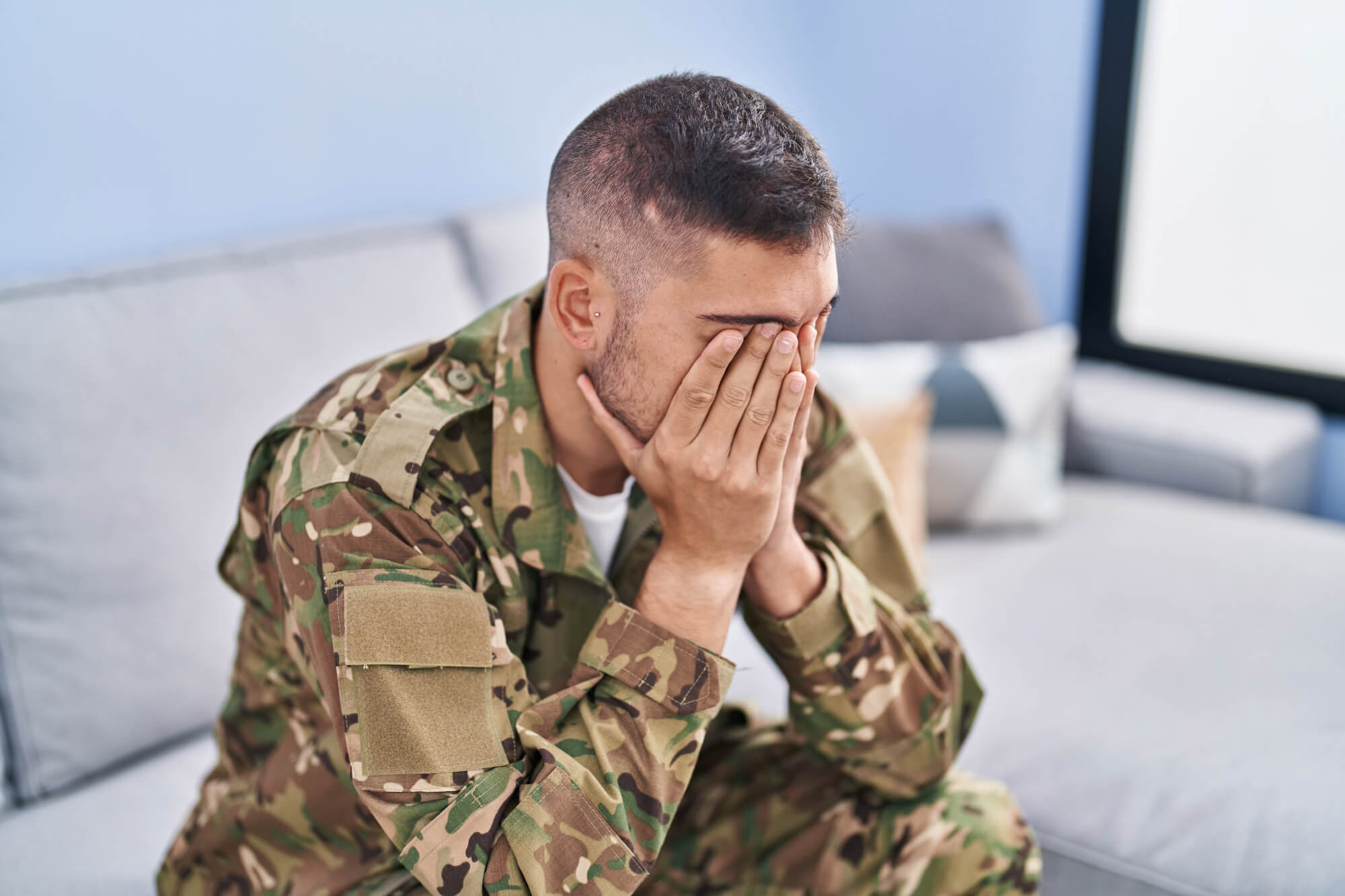 Soldier in camouflage uniform sitting on a couch, covering his face with hands, appearing distressed.