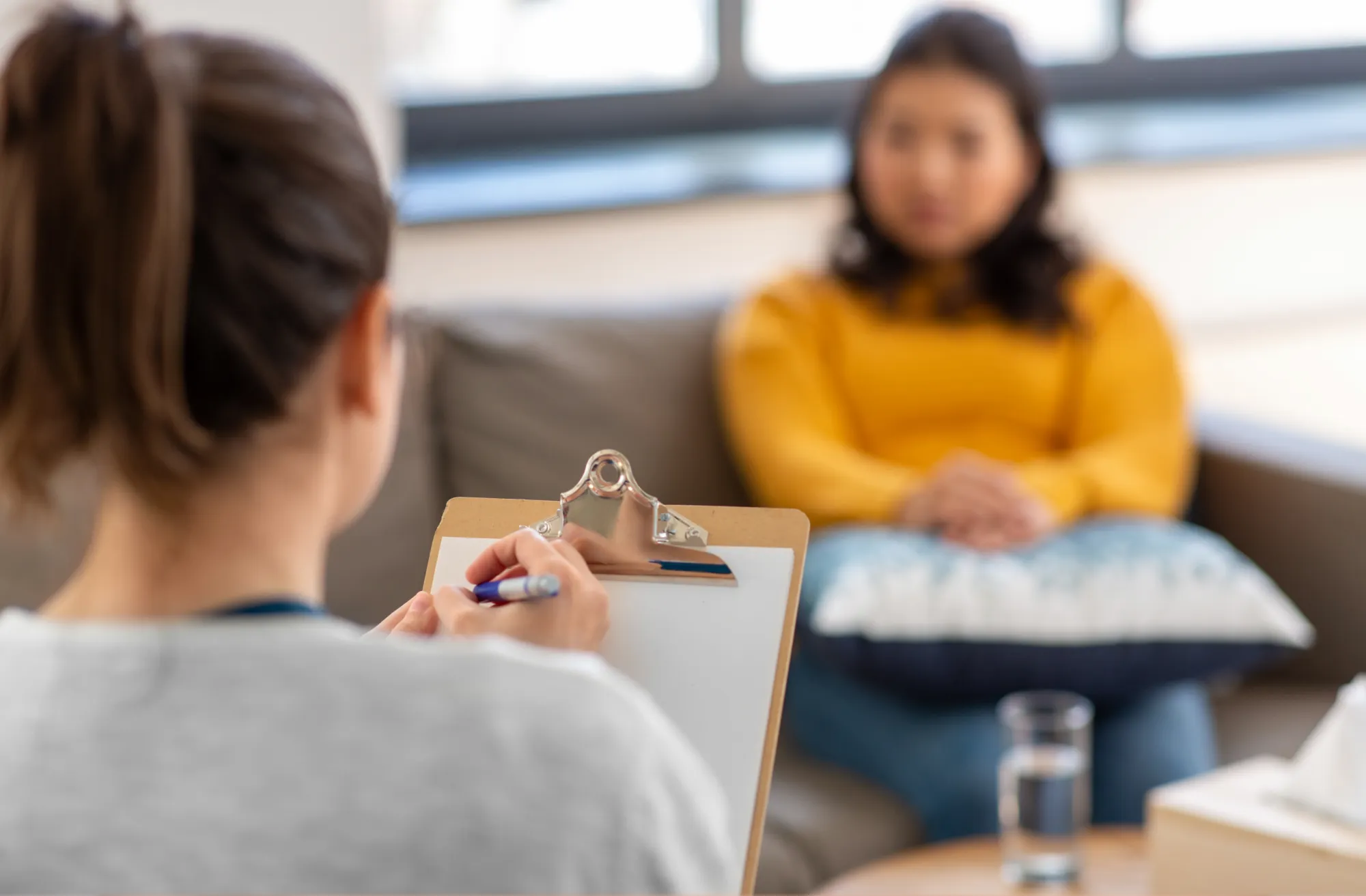 Therapist with clipboard listens to client in yellow sweater during a counseling session in a cozy office.