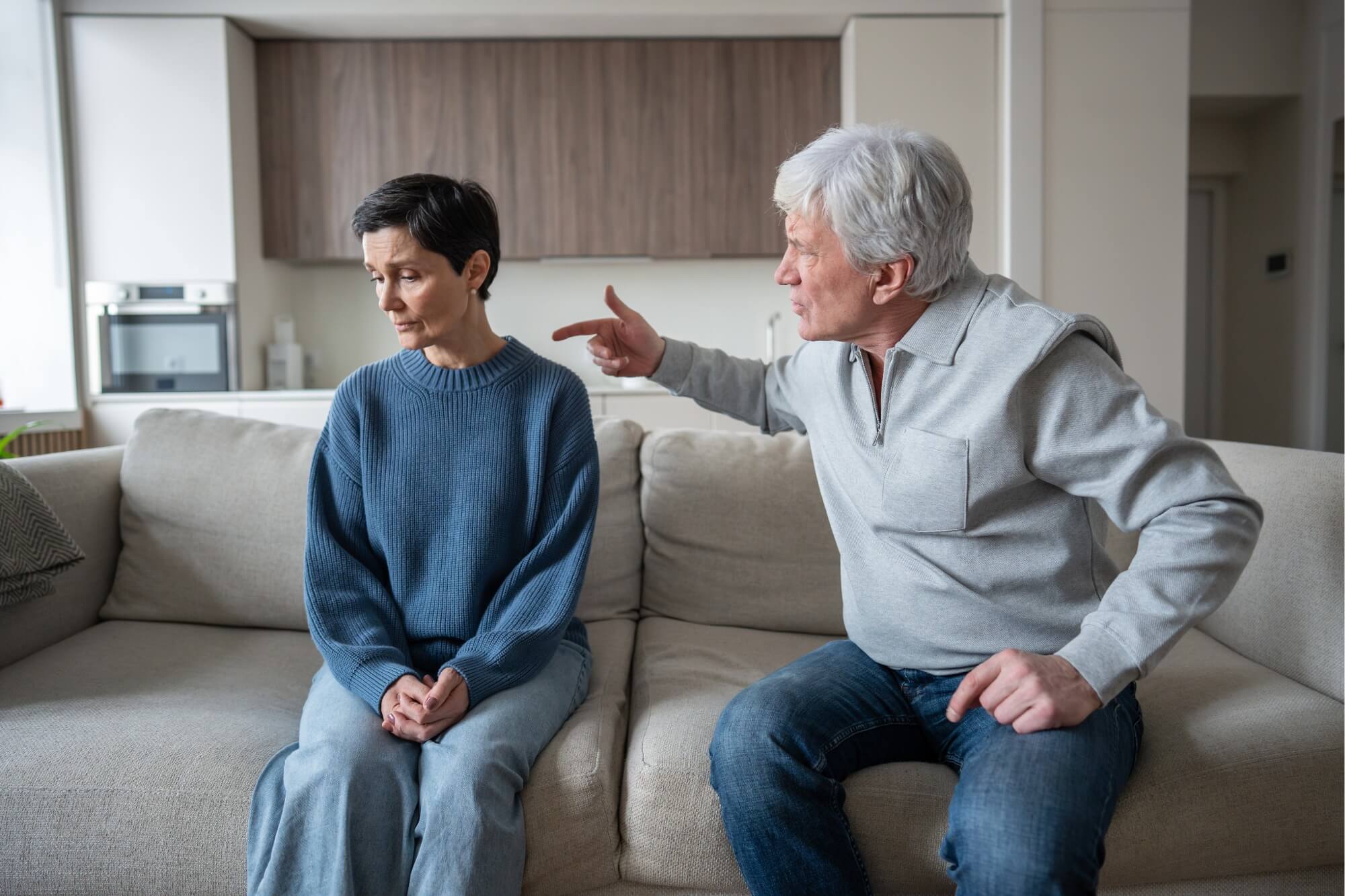 Elderly man pointing finger at sad woman on sofa.