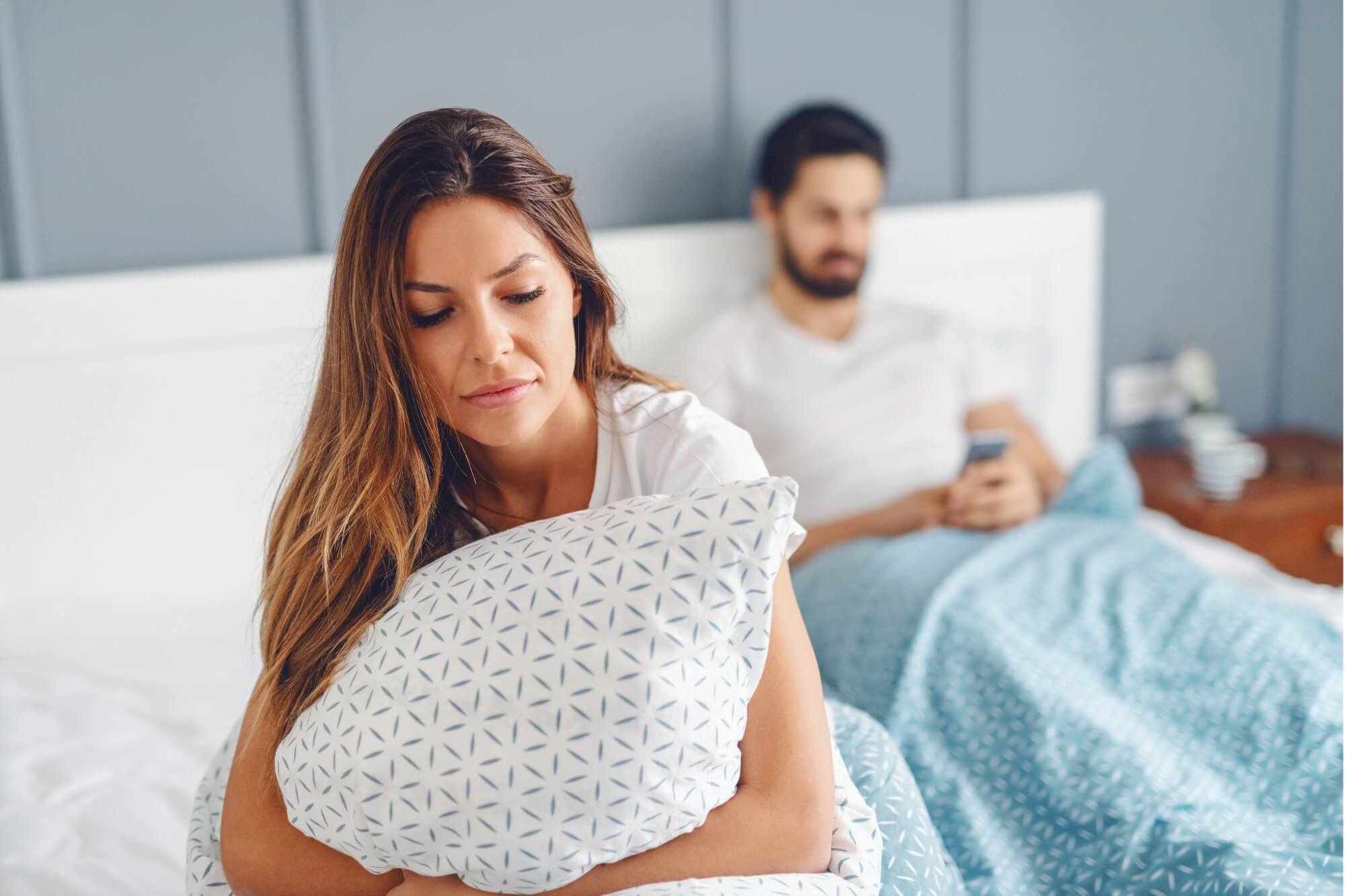 A woman looks upset on a bed with a man using a phone in the background.