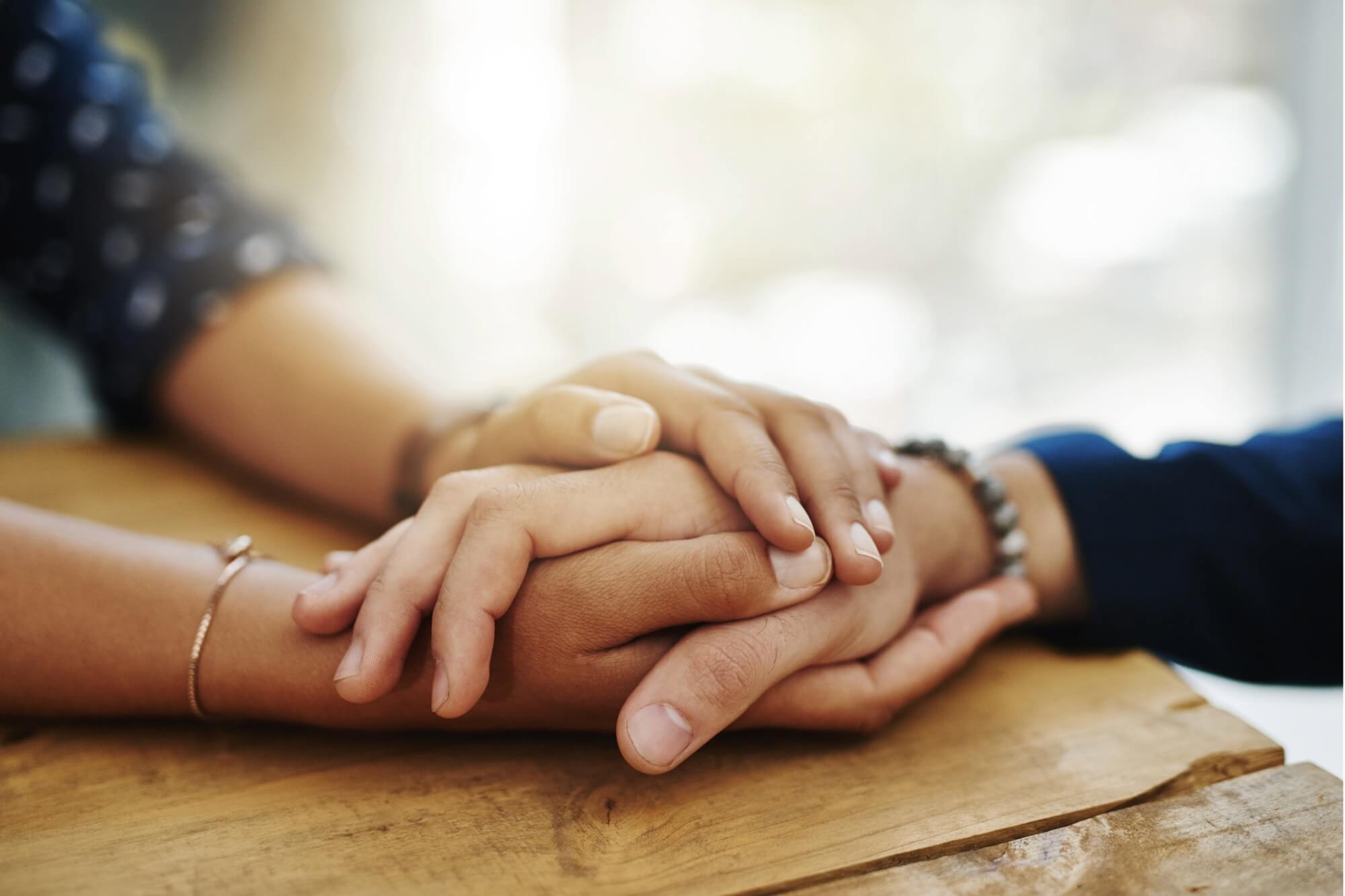 Close-up of two people sitting at a wooden table, holding hands in a comforting gesture, symbolizing support, empathy, and compassion, possibly during a moment of grief or emotional support.