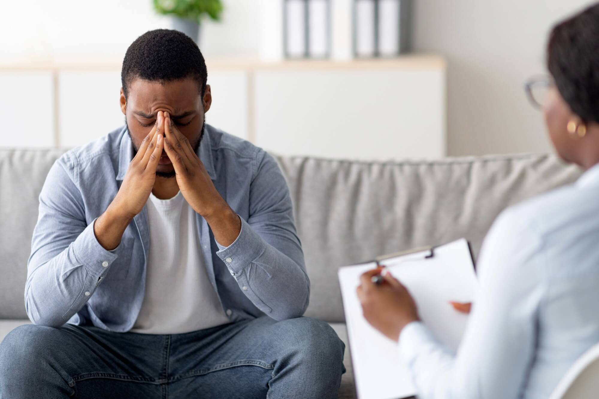 A distressed man sits on a couch with his hands pressed to his face, appearing emotional or deep in thought, while a therapist sits across from him holding a clipboard, taking notes during a counseling session.