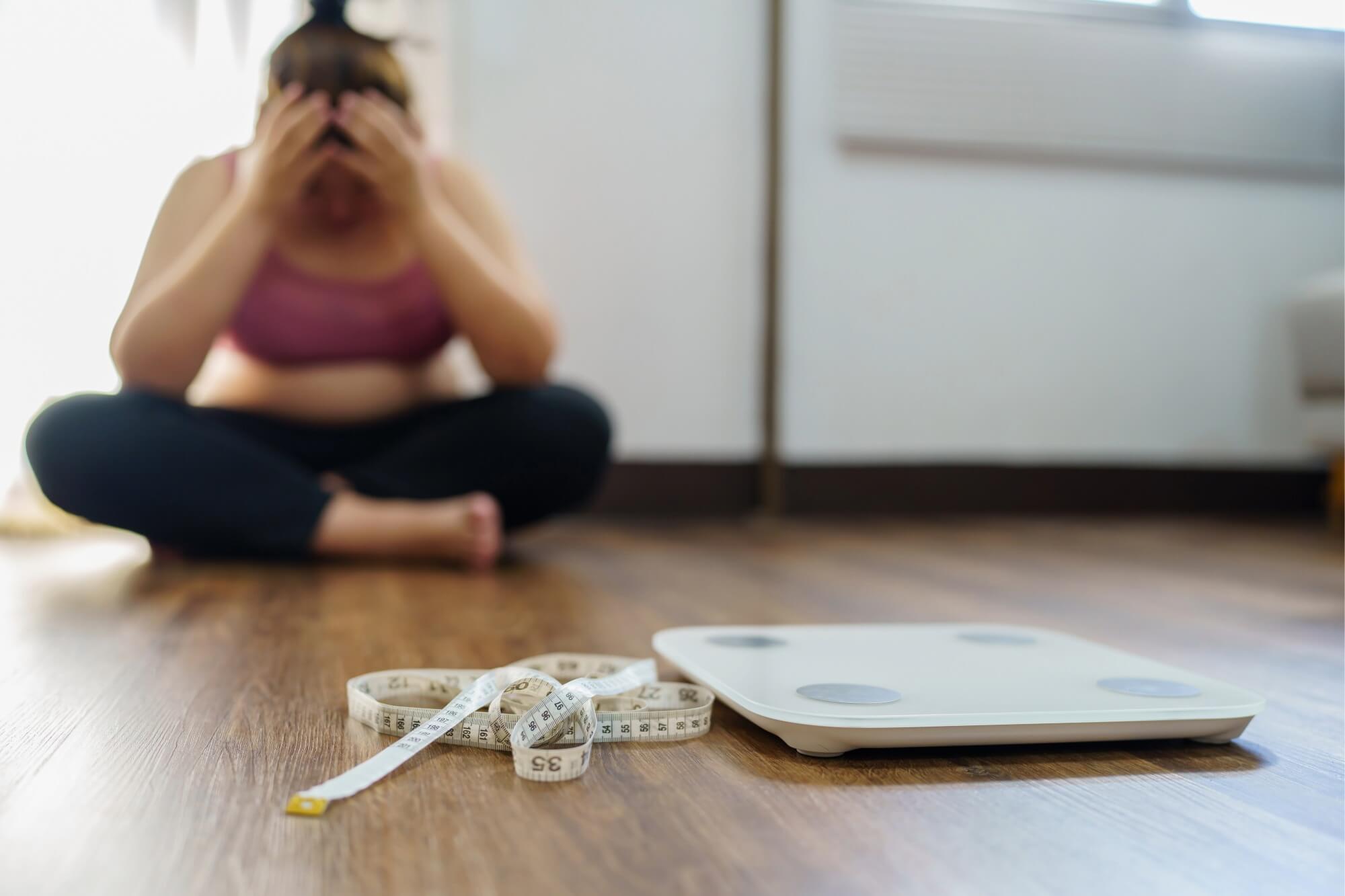 Person sitting on floor, distressed, near a scale and measuring tape in a room.