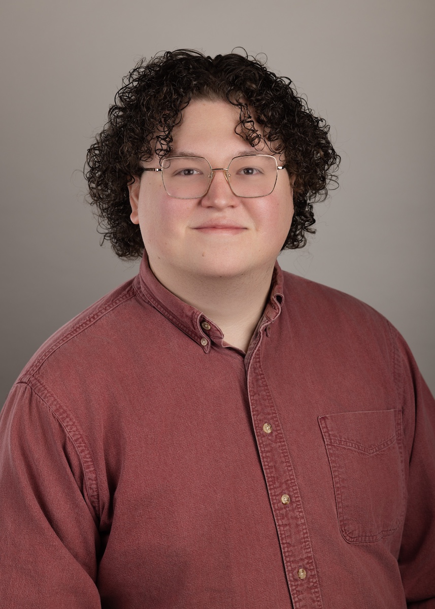 Portrait of a person with curly dark hair and glasses wearing a reddish-brown button-up shirt against a plain gray background.
