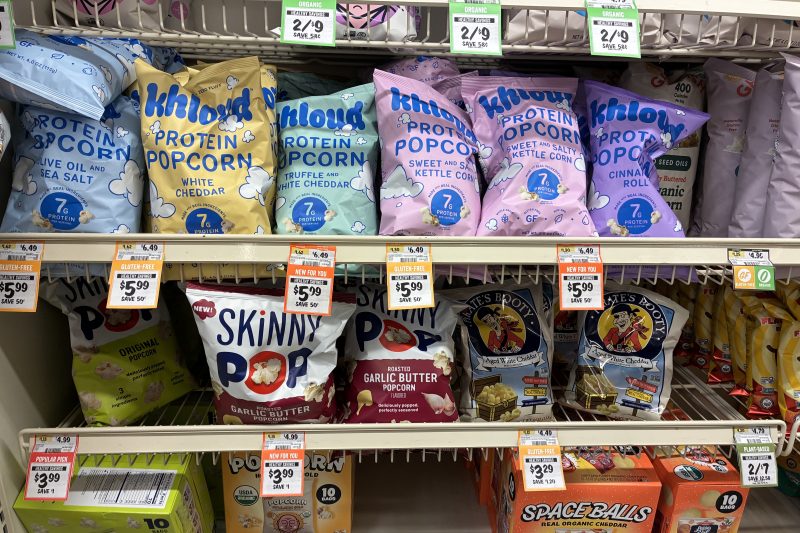 Store shelf displaying various brands and flavors of popcorn, including Khloya protein popcorn in colorful bags on the top shelf and Skinny Pop and Pirate's Booty on the middle shelf with price tags.