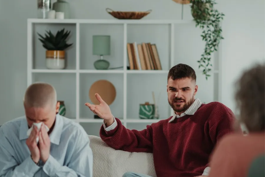 Man in a red sweater talking with expressive hand gestures while a person nearby wipes tears with a tissue during a counseling session.